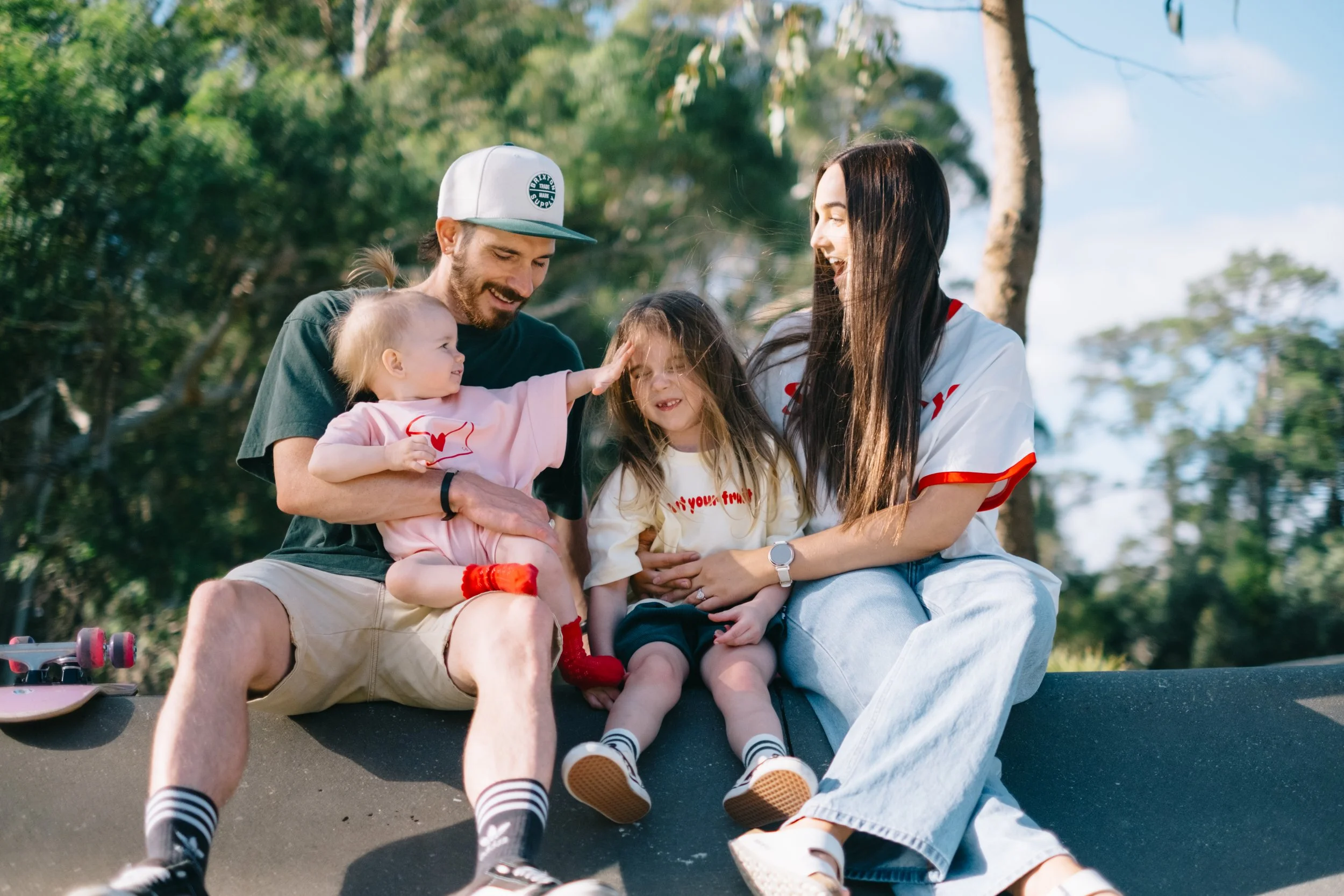 A family of four enjoying a day outdoors in a park, sitting on a skateboard ramp with trees in the background. The father and daughter are playing and smiling, the mother is holding the younger daughter who is wearing a pink outfit, and the older dau