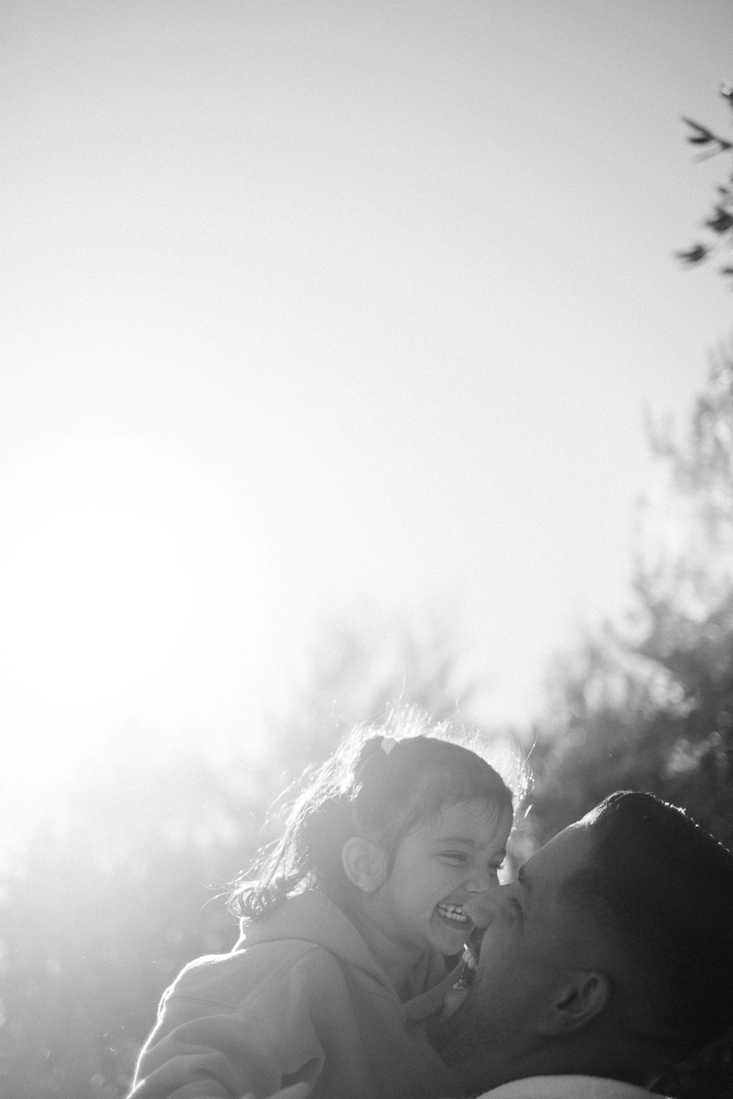 A man holding a young girl, both smiling and looking at each other outdoors in sunlight.