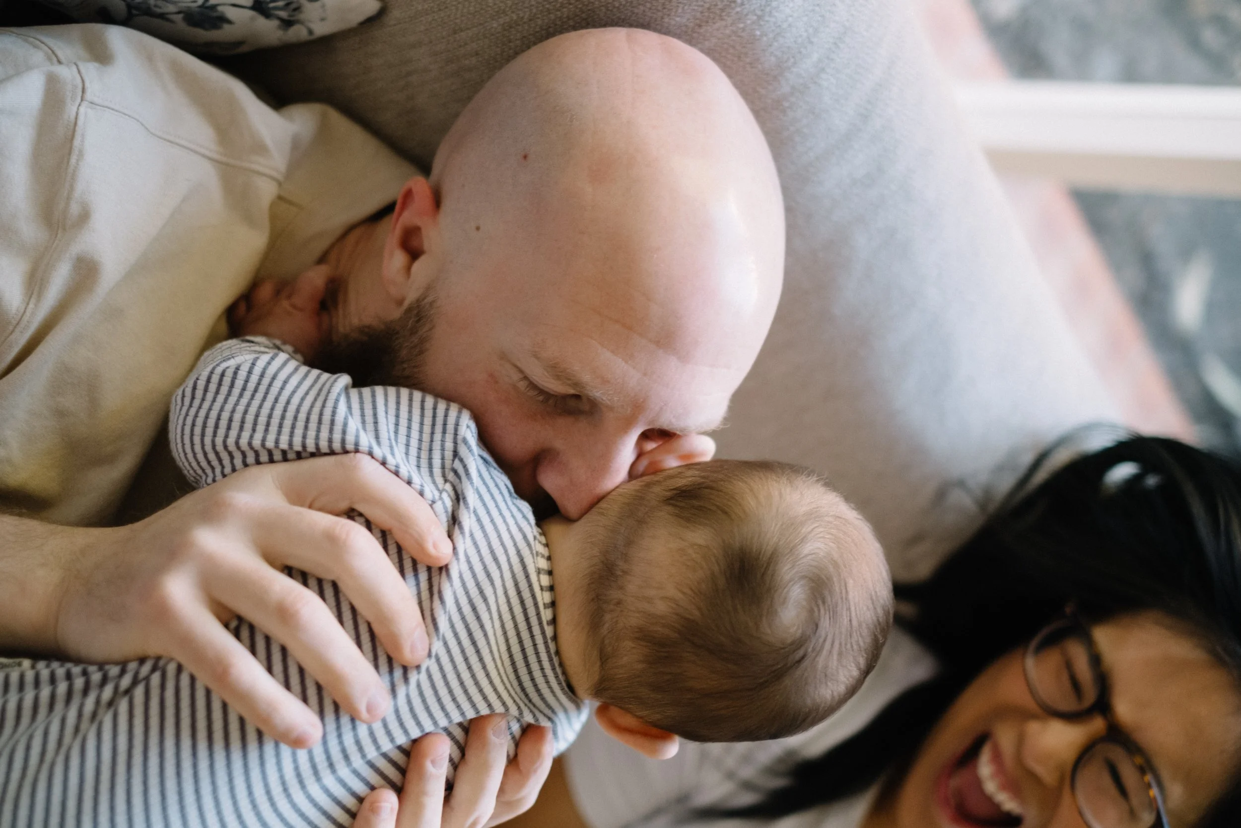 A bald man with a beard and a woman with glasses are lying down on a couch, smiling and looking at a baby, who is wearing a striped shirt.