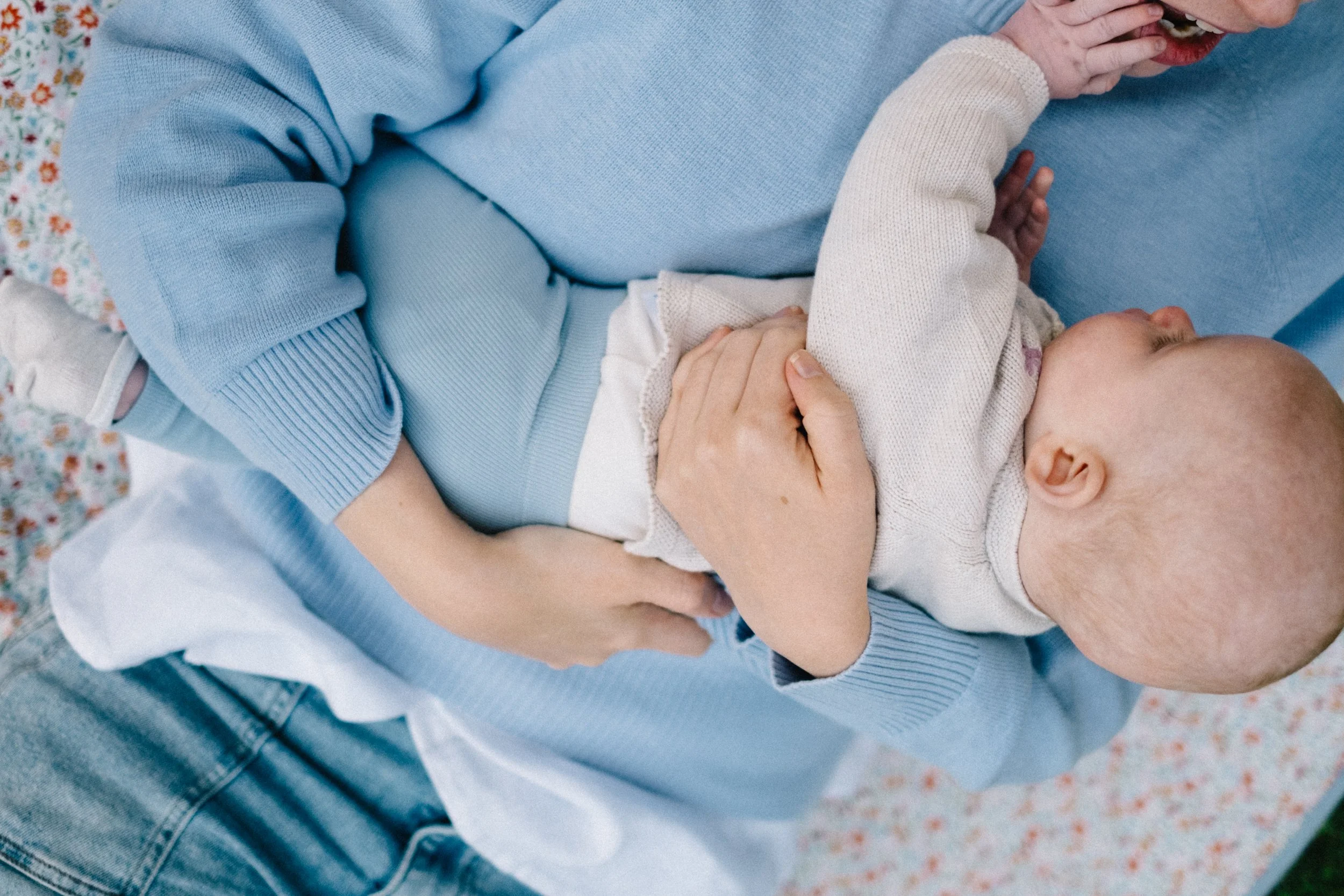 A person in a light blue sweater holding a sleeping baby with a white sweater, lying on a blanket