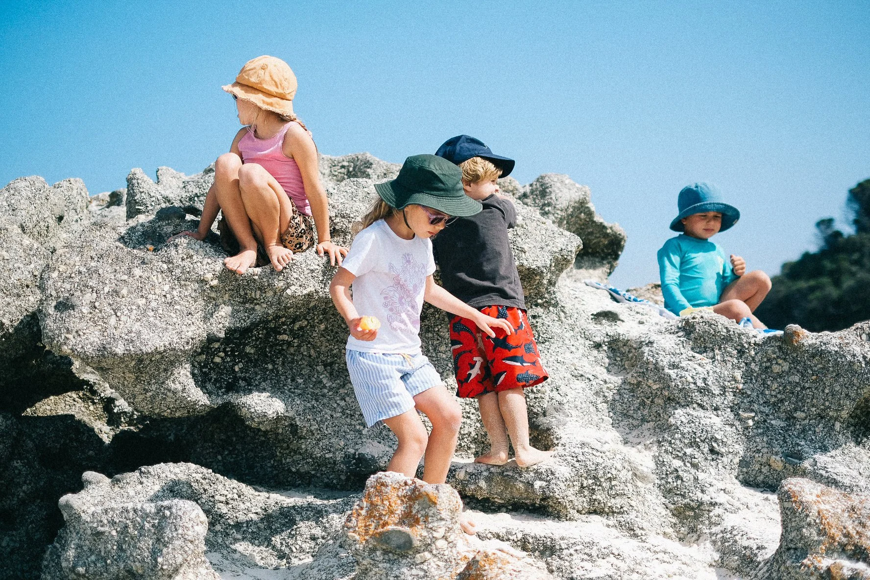 Group of children playing on rocky beach, some sitting on rocks and others walking, under a clear blue sky.