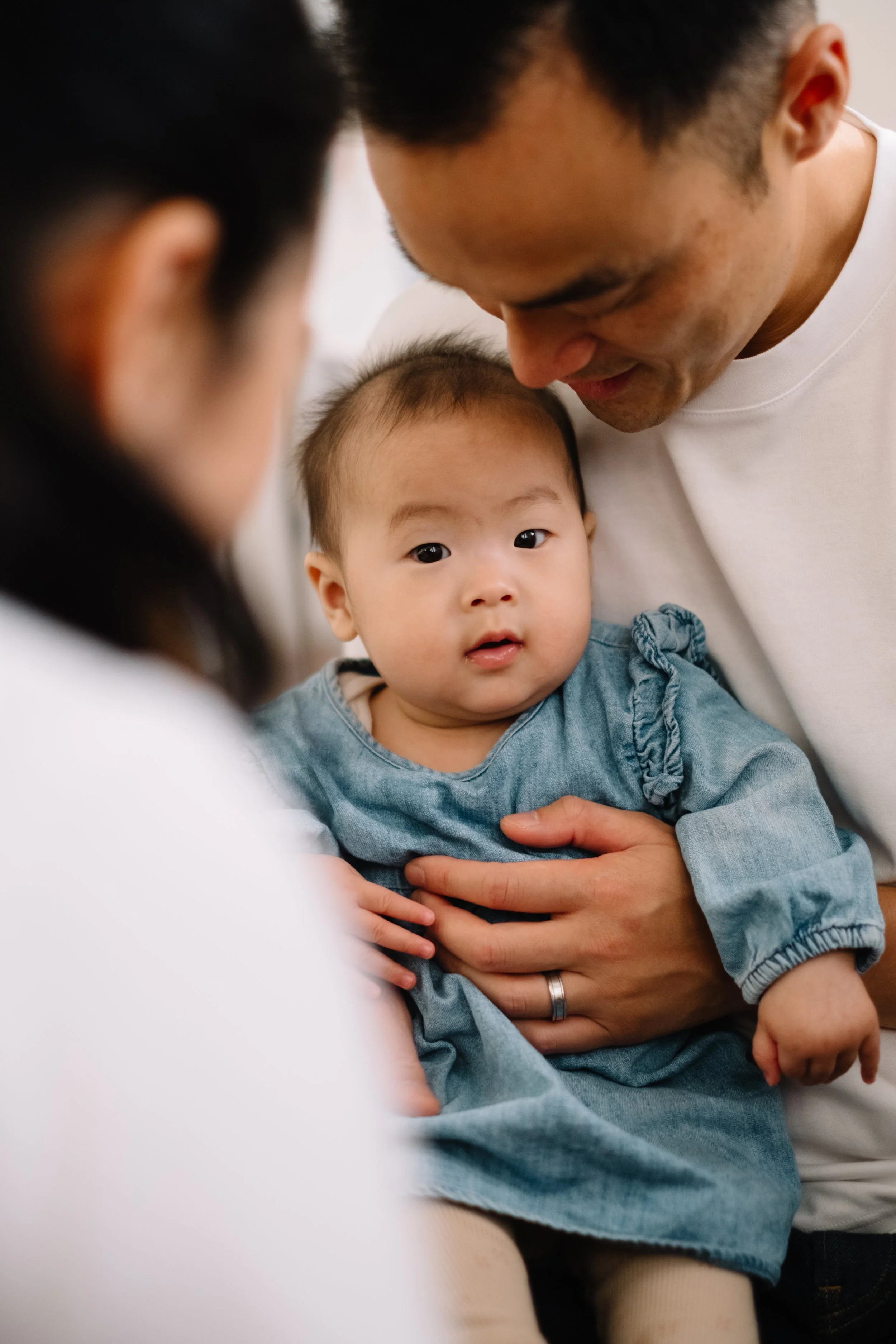 A young Asian child in a blue dress being held and gently touched by an adult man, with another adult lady partially visible in the foreground. The child and adults are close together in a warm, intimate setting.