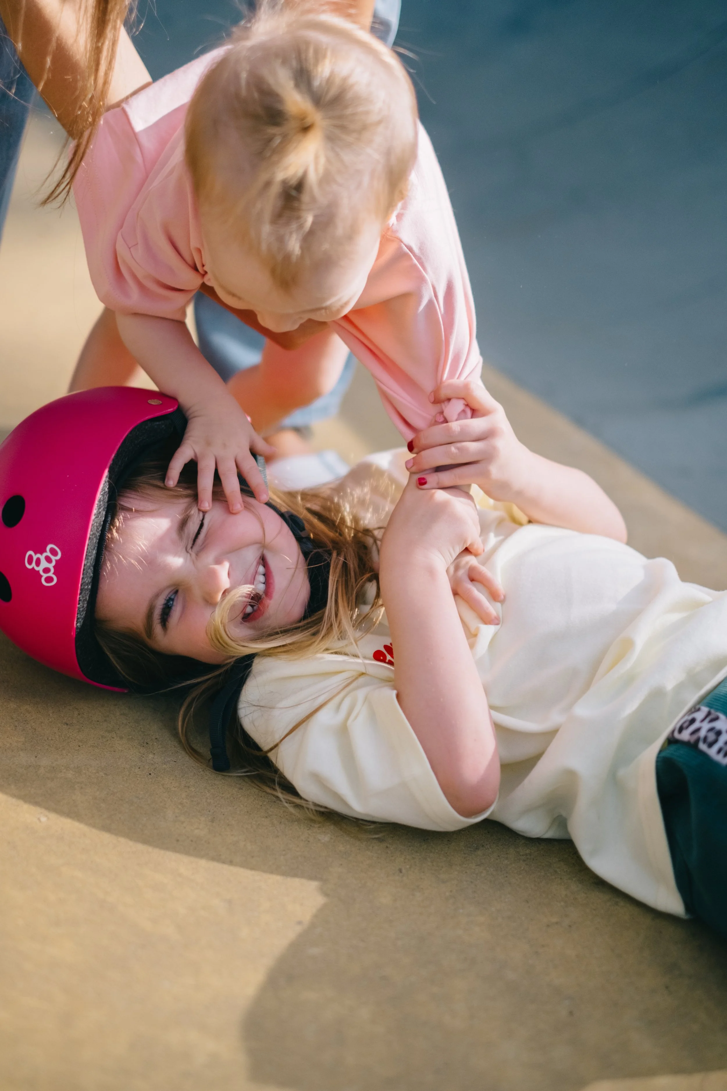 Two young girls play fight on the ground; one girl is lying down wearing a pink helmet and a cream-colored shirt, smiling, while the other girl is leaning over her, pulling her arm and touching her forehead with her hand.