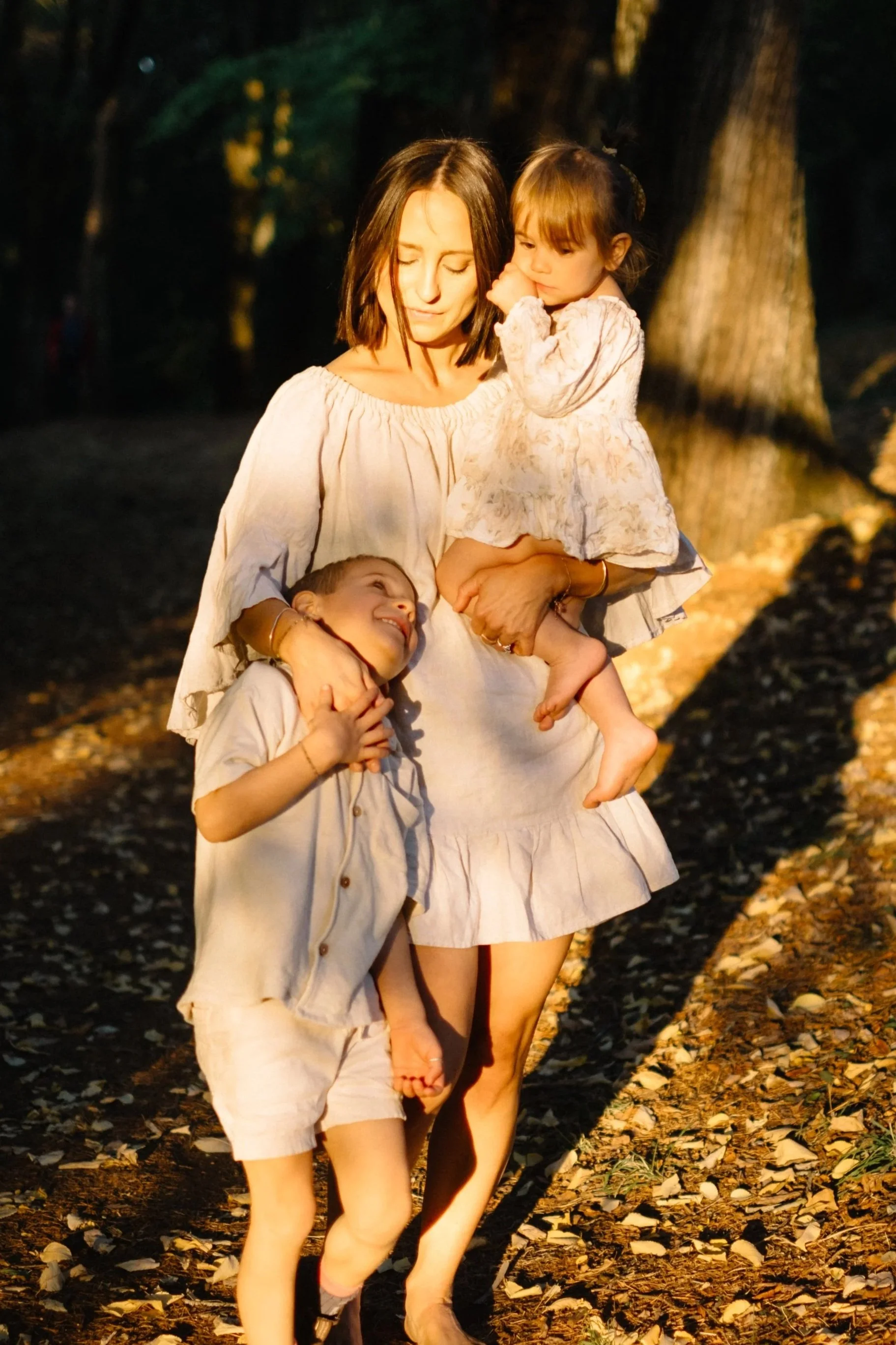 A woman walking outdoors with two children, one in her arms and one holding her hand, surrounded by trees and fallen leaves.