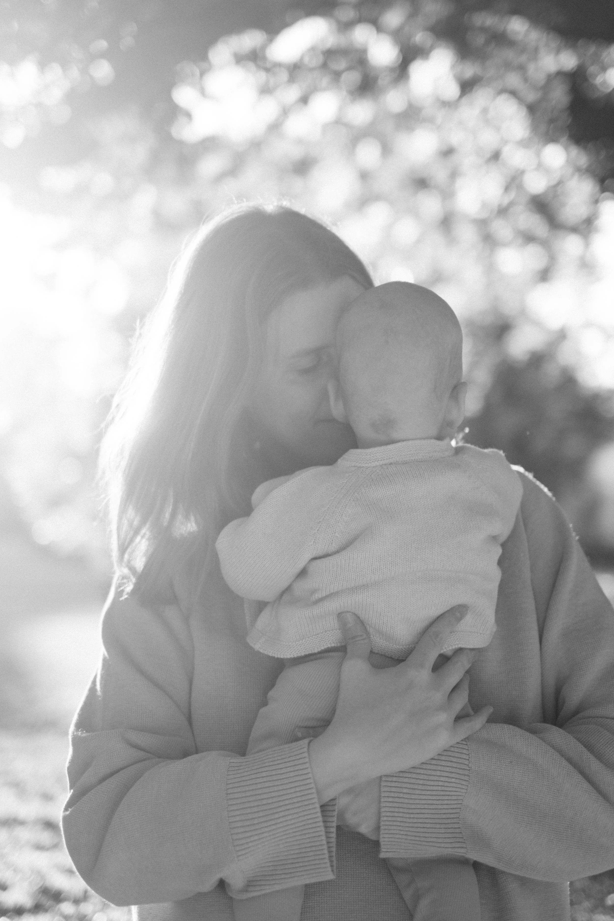 A woman holding a young child outdoors with sunlight filtering through trees in the background.