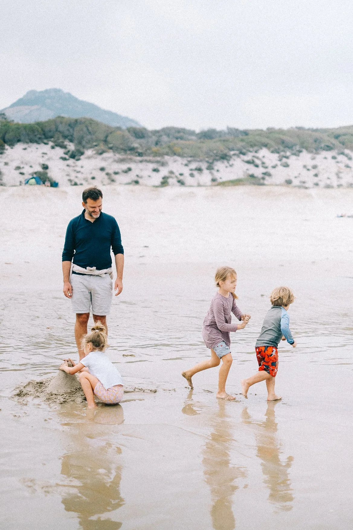 A man playing with three children at the beach, building a sandcastle and walking in shallow water.
