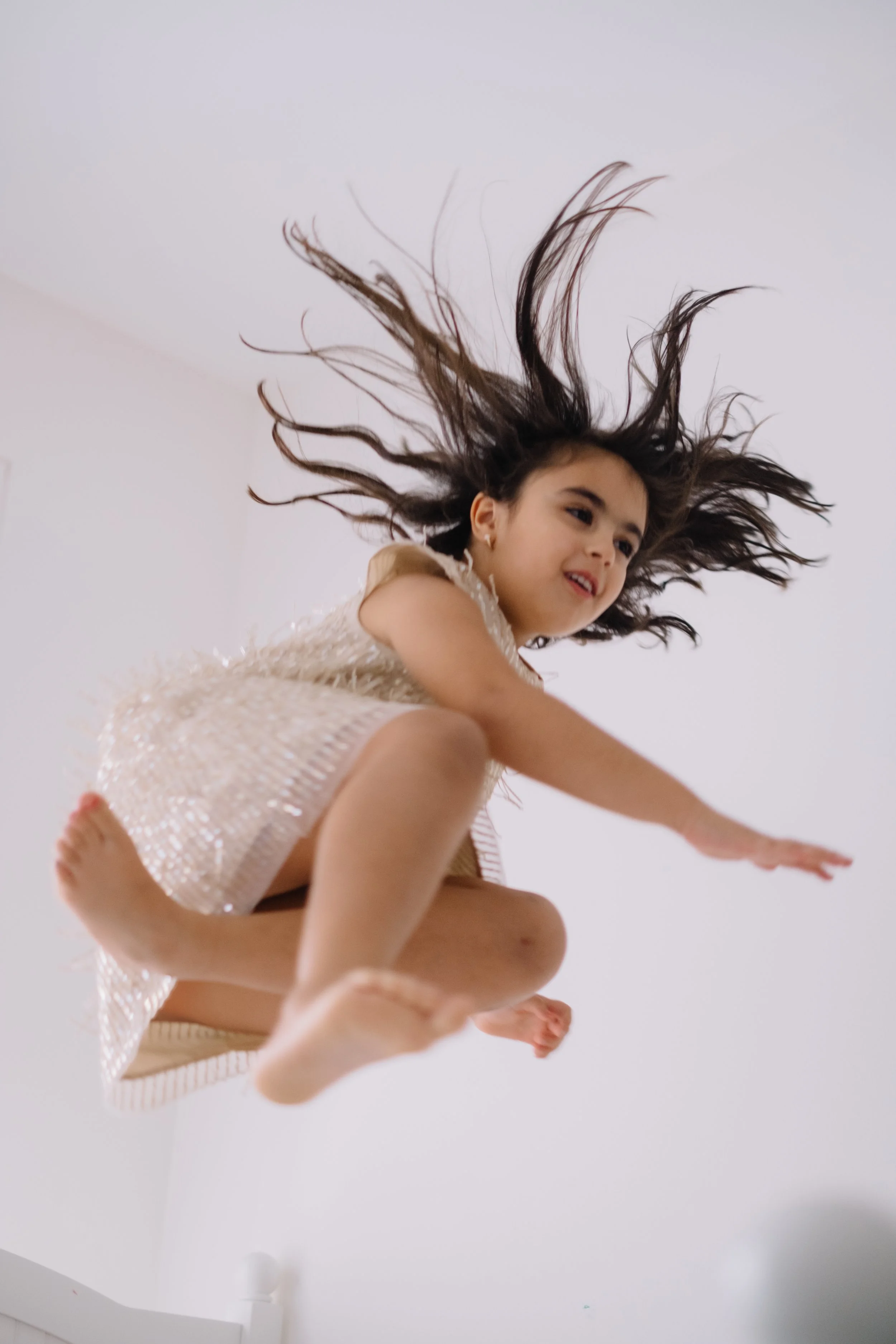 Young girl jumping in the air with hair flowing, wearing a light-colored dress, against a plain white background.