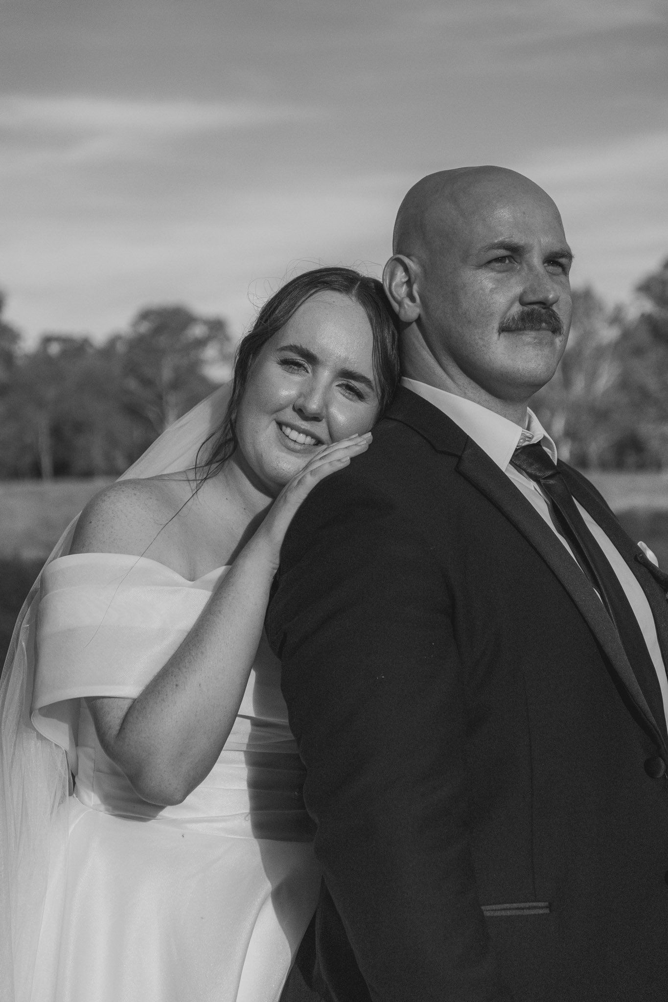A black and white photo of a bride and groom outdoors. The bride is smiling and resting her head on the groom's shoulder, with her hand on his back. The groom, dressed in a suit, looks away from the camera.