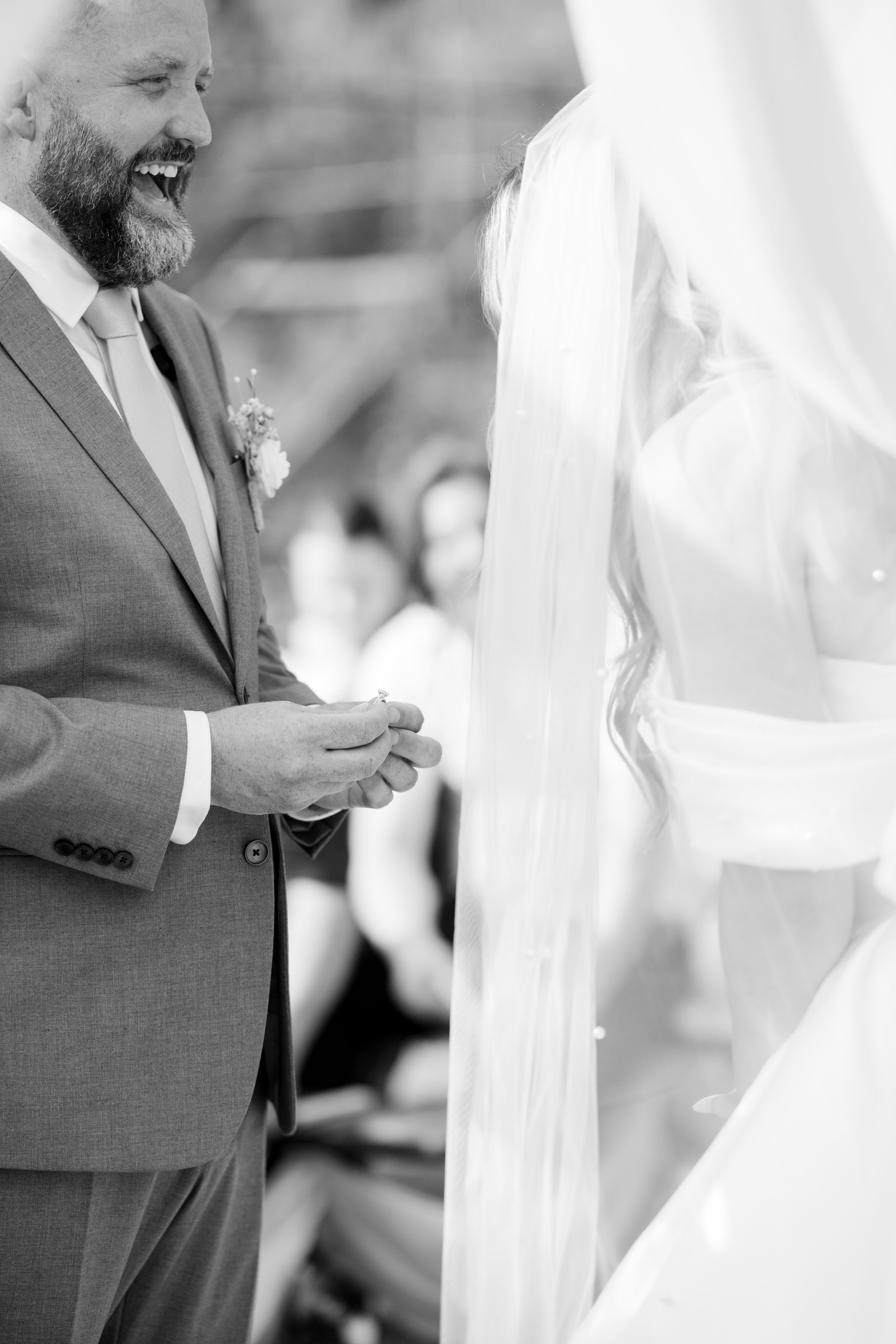 A black and white photo of a wedding ceremony showing a man in a suit smiling and looking at a woman in a wedding dress with a veil.