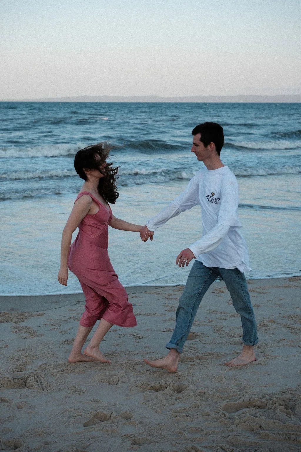 A man and woman holding hands and dancing on the beach near the ocean, with waves in the background and sandy ground under their feet.