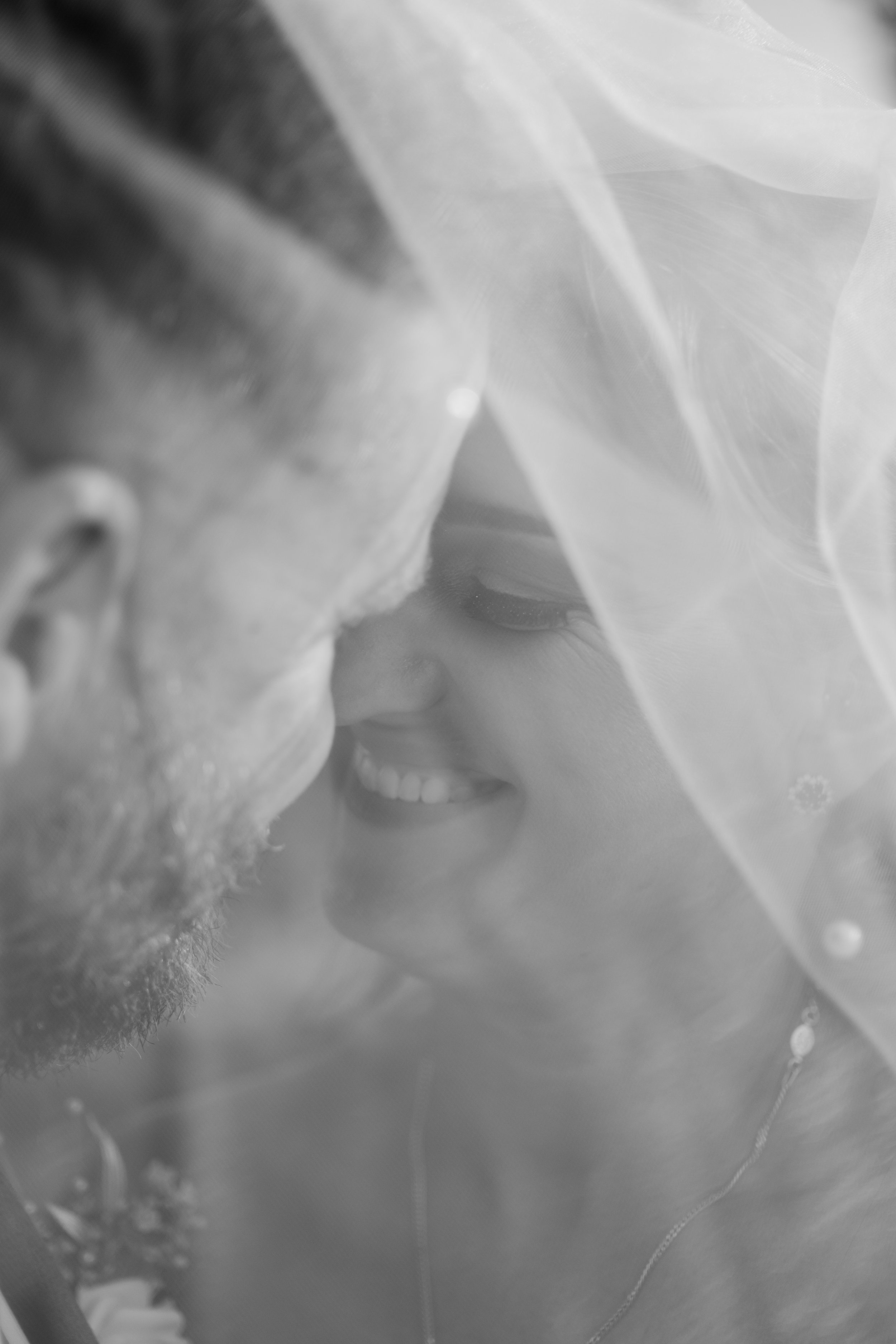 A close-up black and white photo of a couple smiling and leaning close together, with a veil partially covering their faces.