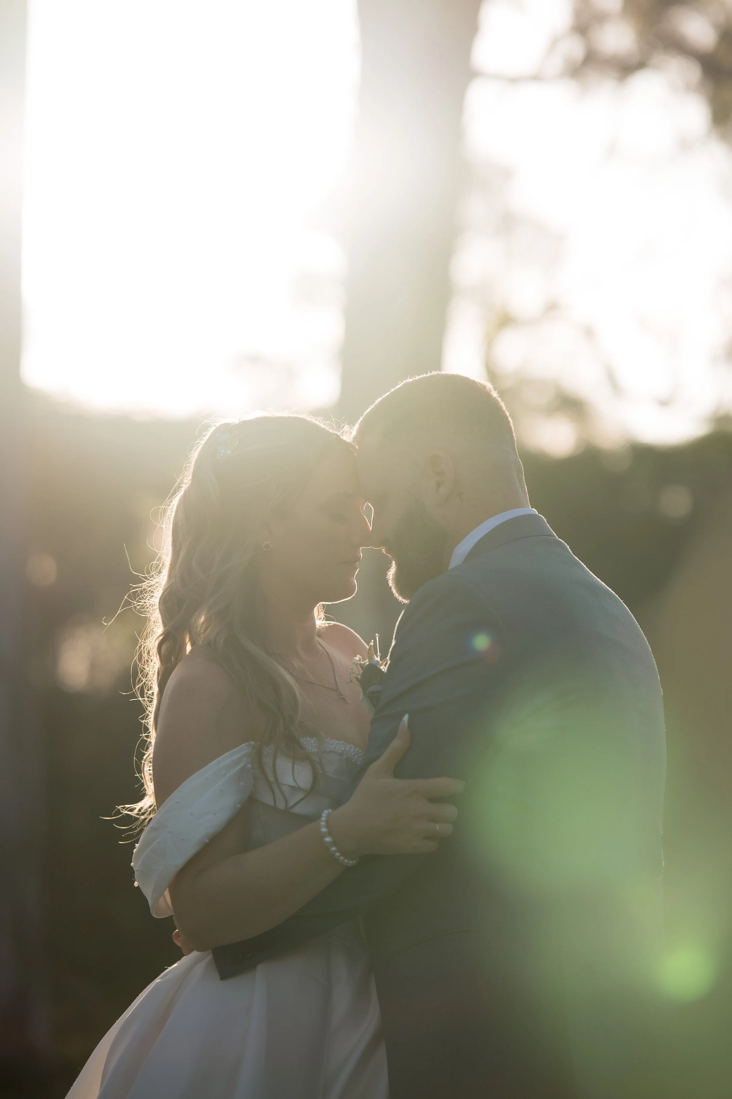 A couple in wedding attire share a romantic moment outdoors, backlit by sunlight, with their foreheads touching and eyes closed.