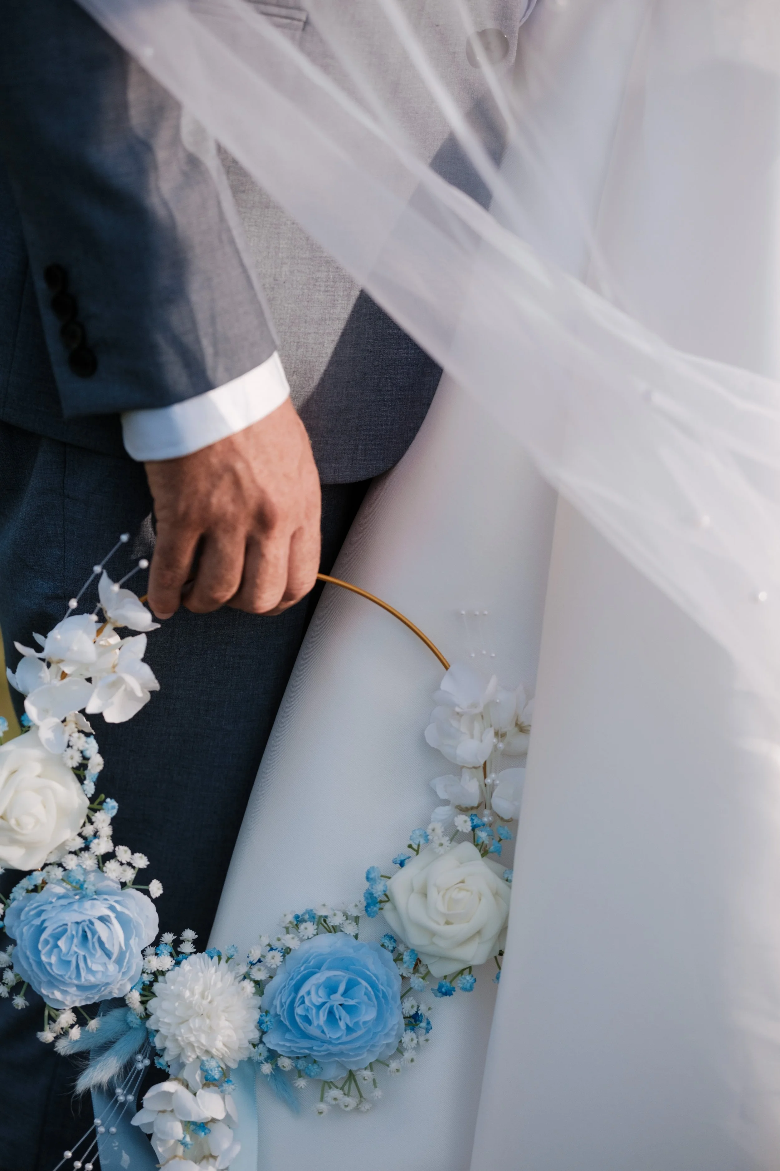 Close-up of a groom in a gray suit holding a wedding bouquet of white and blue flowers, with part of his hand and suit visible.