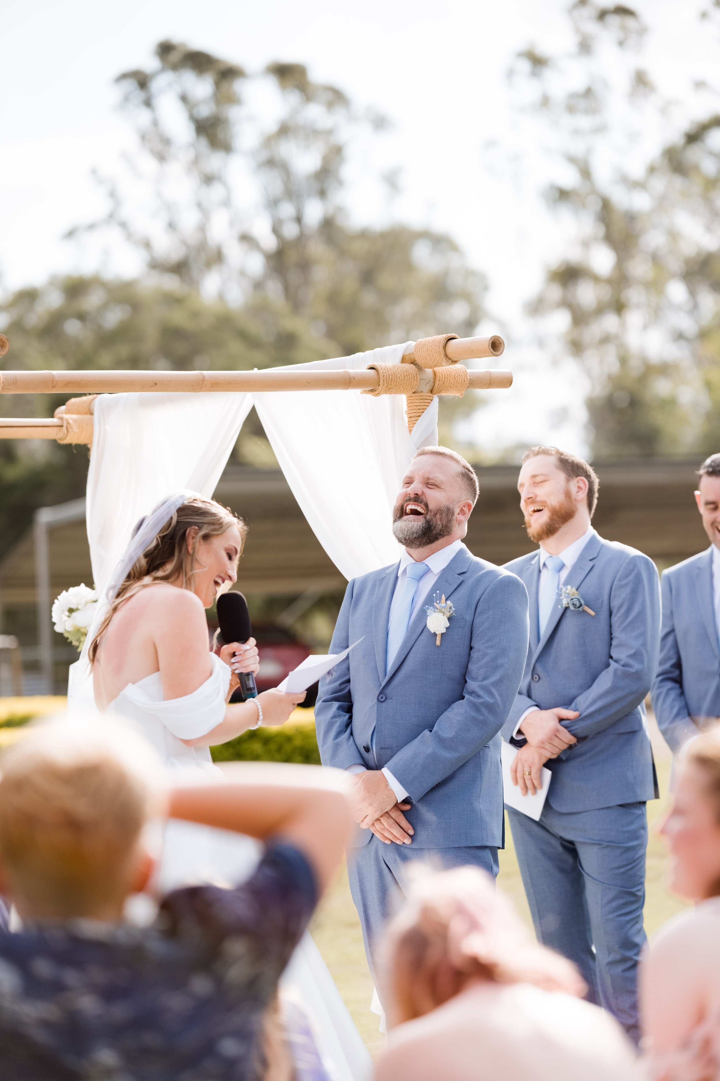 Bride and groom exchanging vows at an outdoor wedding ceremony, with bridesmaids and groomsmen, under a wooden arch with white drapery and greenery, on a sunny day.