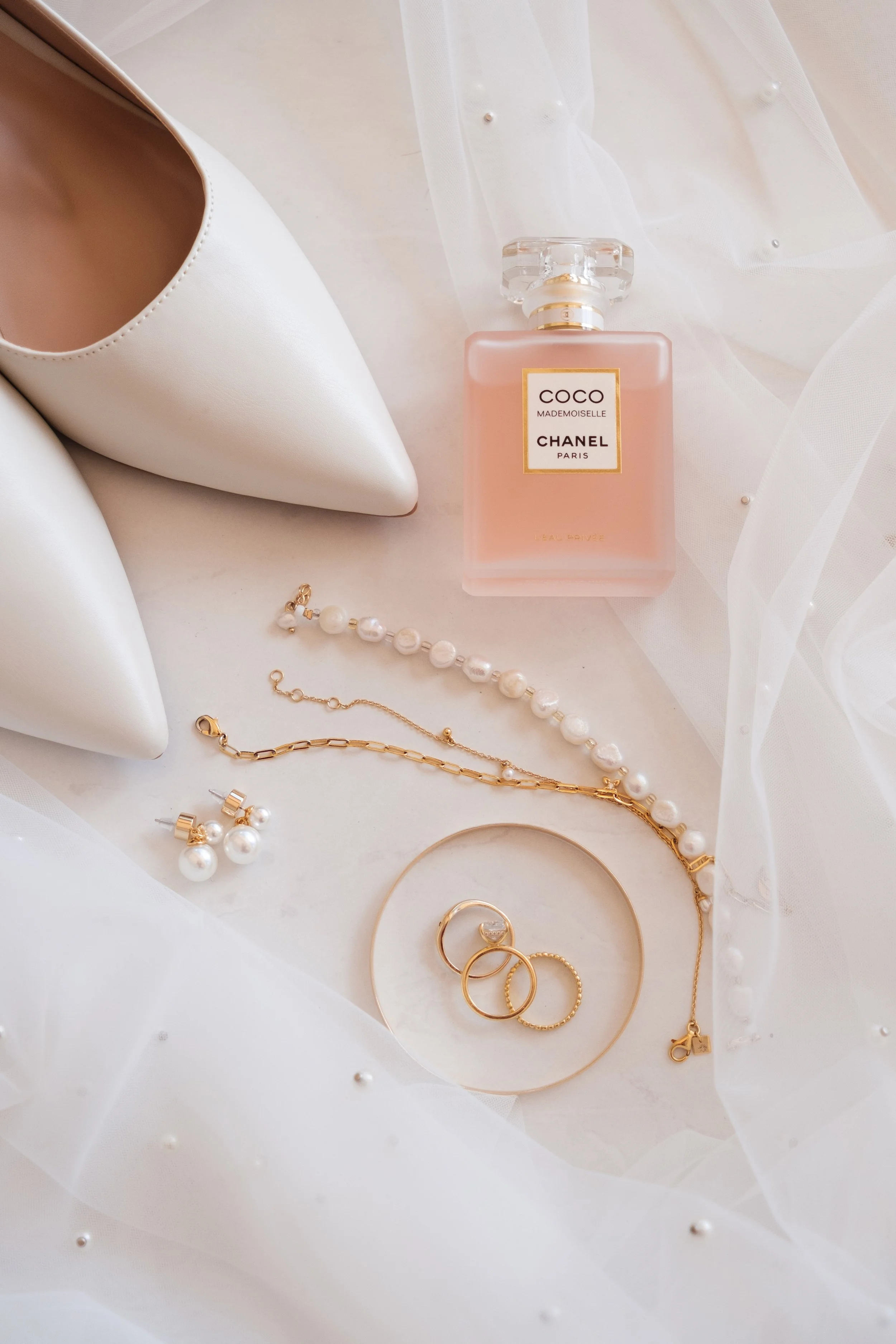 A pair of white pointed-toe high heels, a bottle of Chanel Coco Mademoiselle perfume, pearl jewelry including a necklace, earrings, rings, and a gold bracelet, all arranged on a white surface with delicate white fabric with pearl accents.