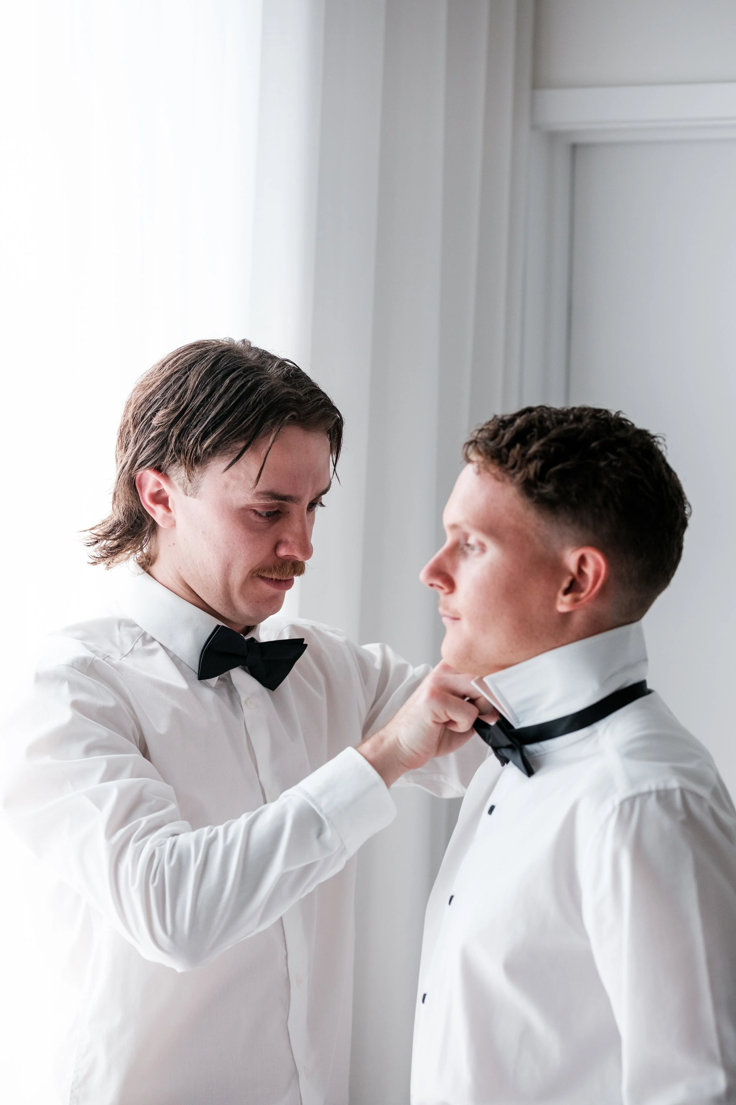 A man adjusts another man's black bow tie in a well-lit room, both dressed in white shirt and black bow tie, preparing for a formal event.