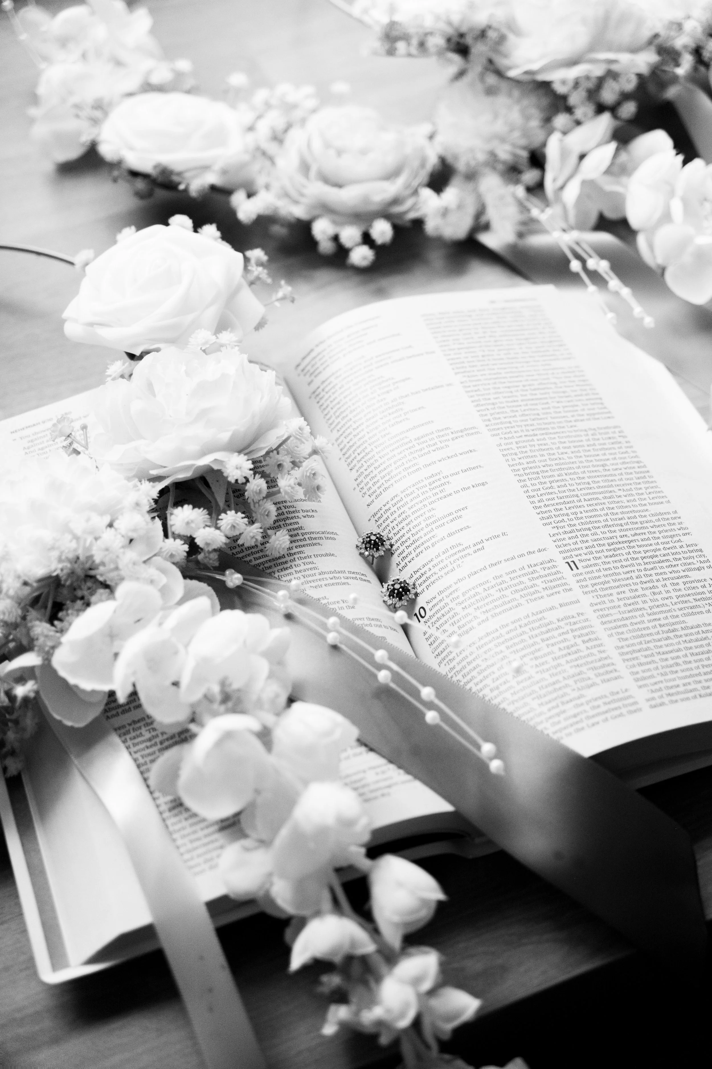 Black and white photo of an open Bible with flower arrangements, including roses and other blooms, placed on a wooden table.