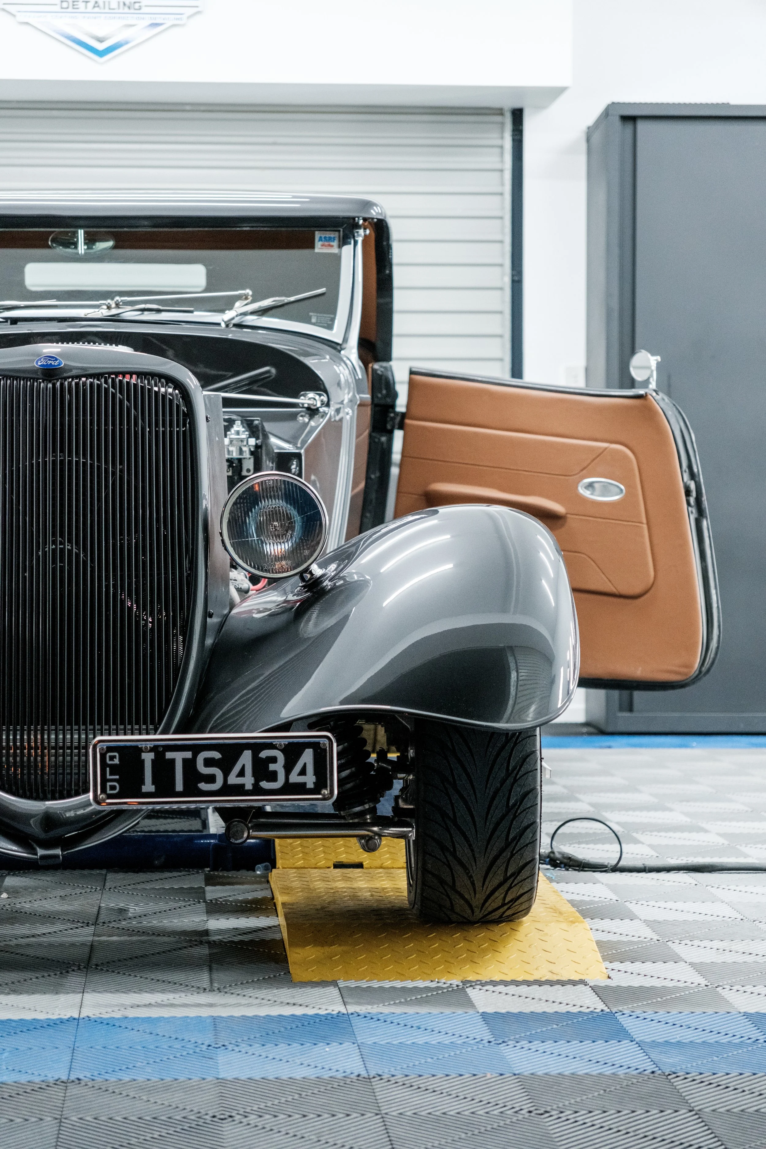 Front view of a vintage black Ford car with a tan door open, showing details of the grille, headlight, and wheel, parked indoors on a black, gray, and blue checkered floor.