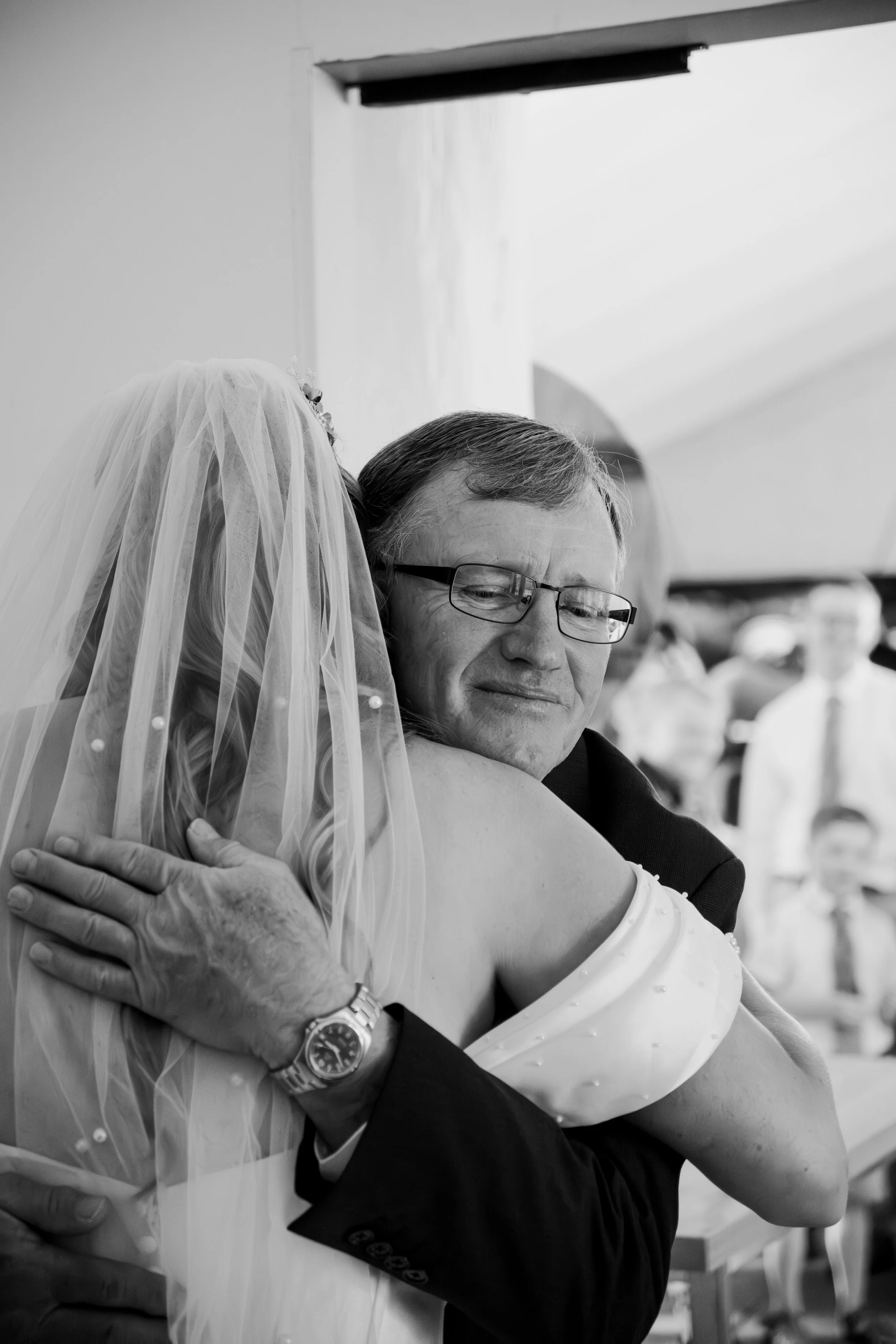 A man and a woman, dressed for a wedding, hugging each other affectionately. The woman is wearing a veil and the man is wearing glasses and a suit. The setting appears to be a wedding celebration.