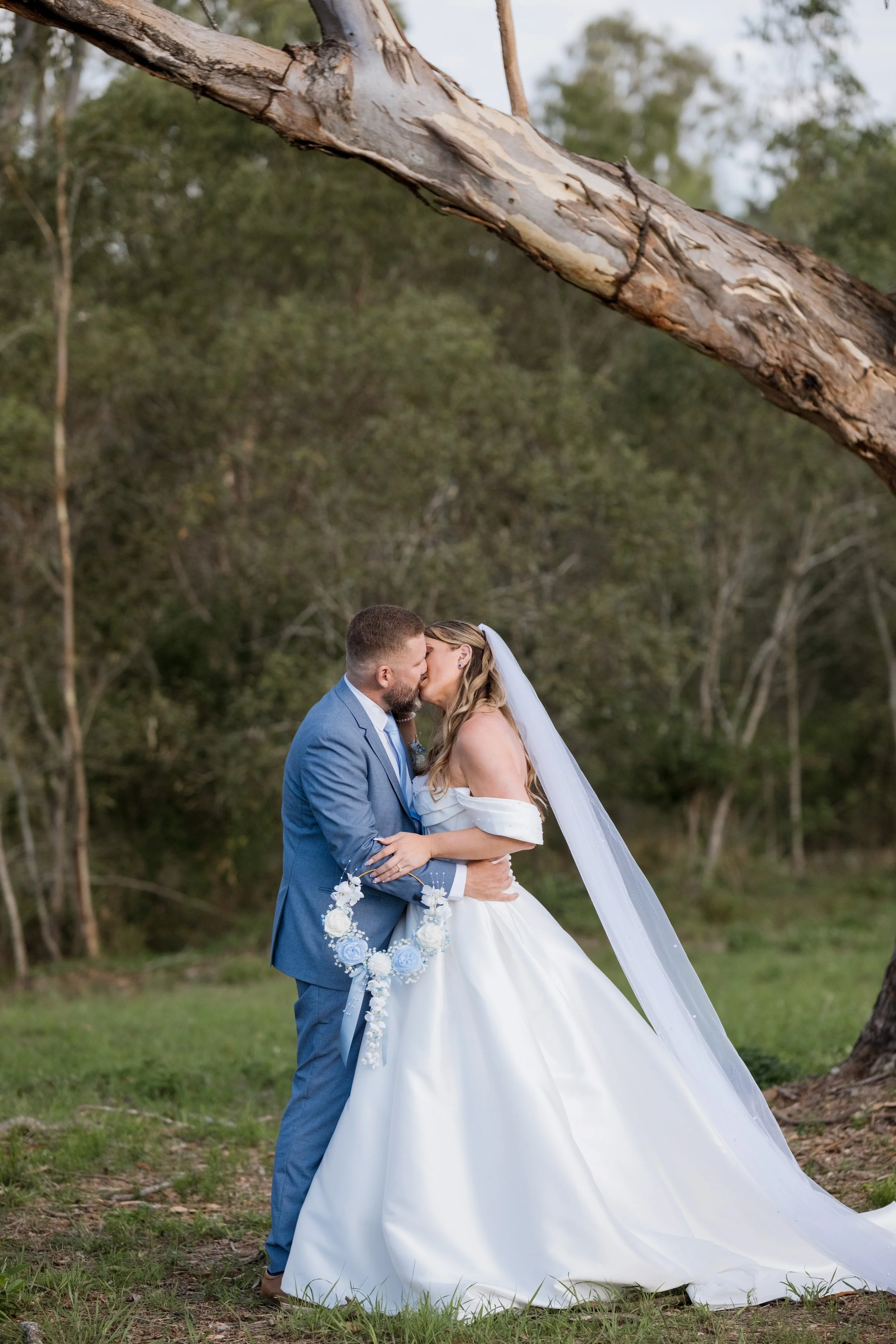A bride and groom kissing outdoors during their wedding, with a large fallen tree and green forest in the background.