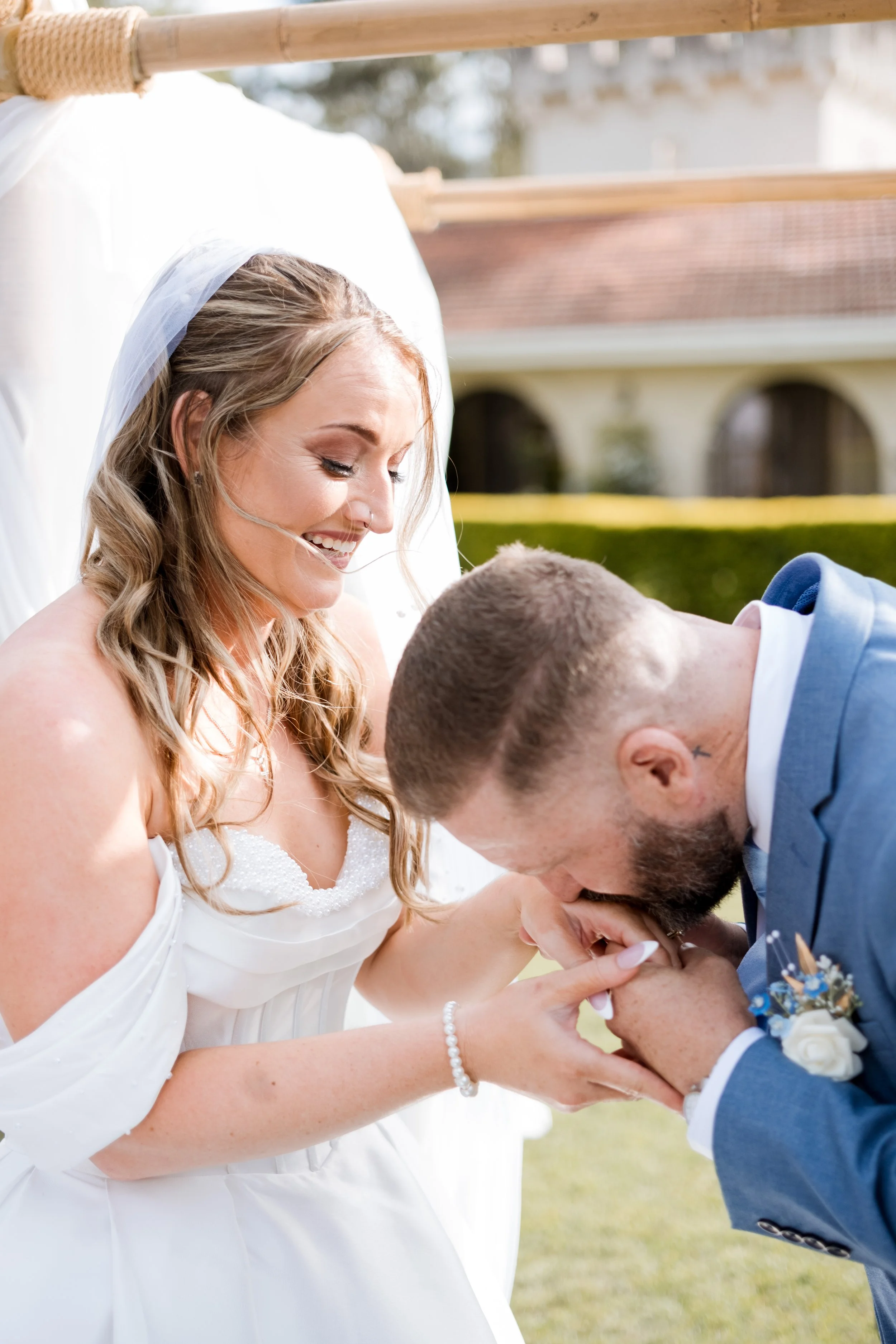 A woman in a wedding dress and veil smiles as she holds the hand of a man in a blue suit who is kissing her hand during an outdoor wedding ceremony.