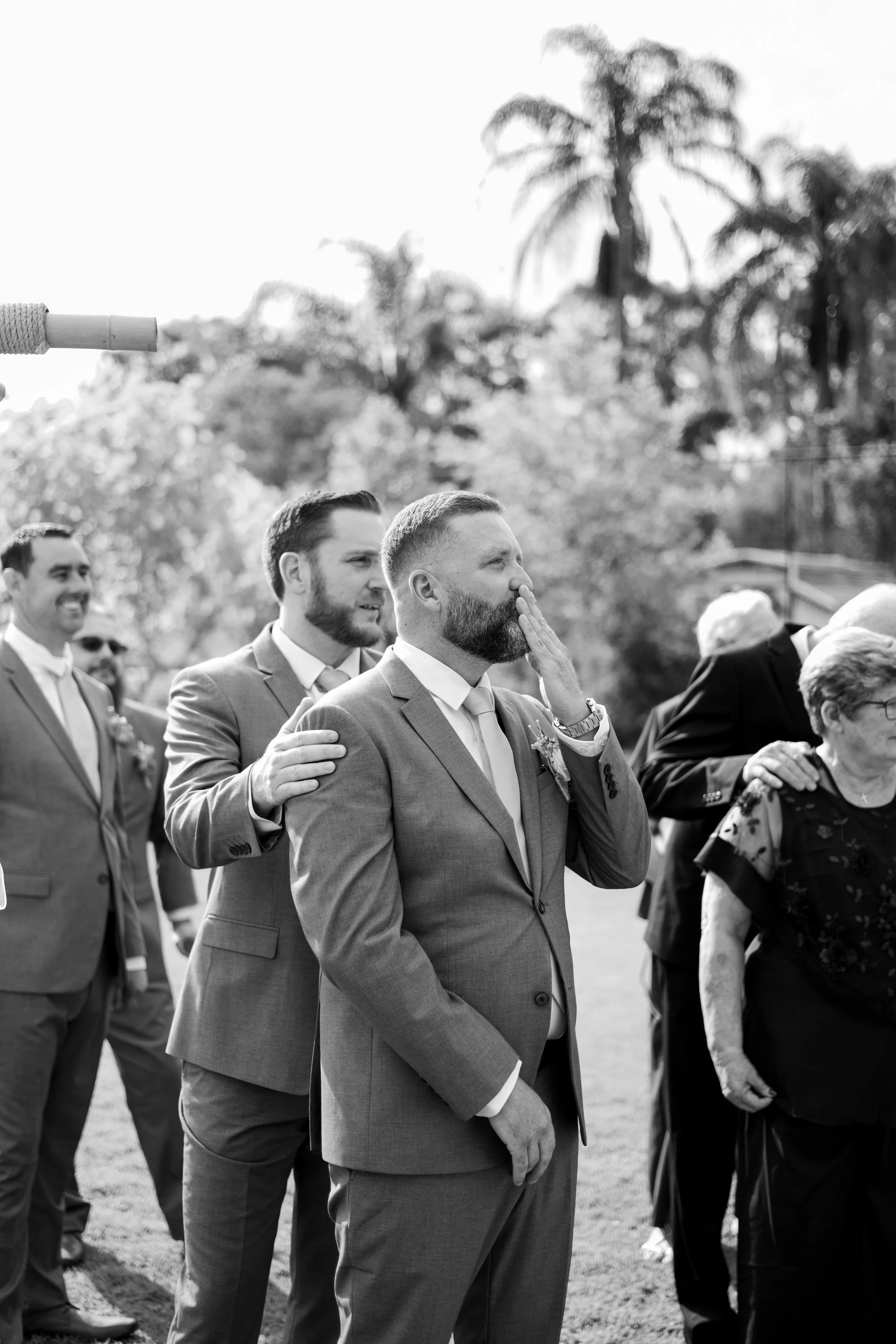 Black and white photo of men in suits, standing in a line outdoors at a wedding or special event, with a man in the foreground touching his face and others smiling or with their hands on each other's shoulders, trees and a cloudy sky in background.