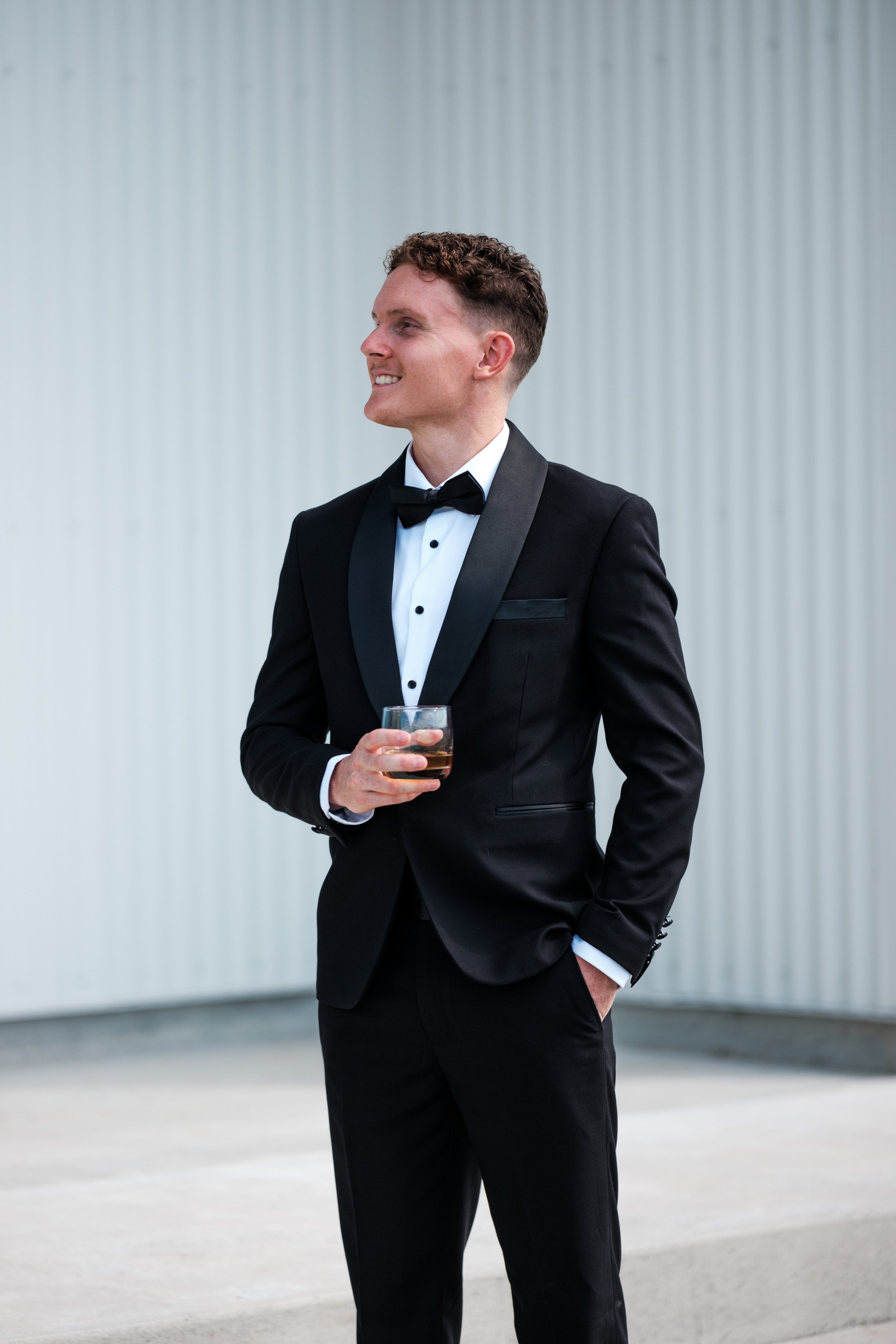 Man in a tuxedo holding a glass of whiskey, standing outdoors against a white corrugated metal wall, smiling and looking to the side.