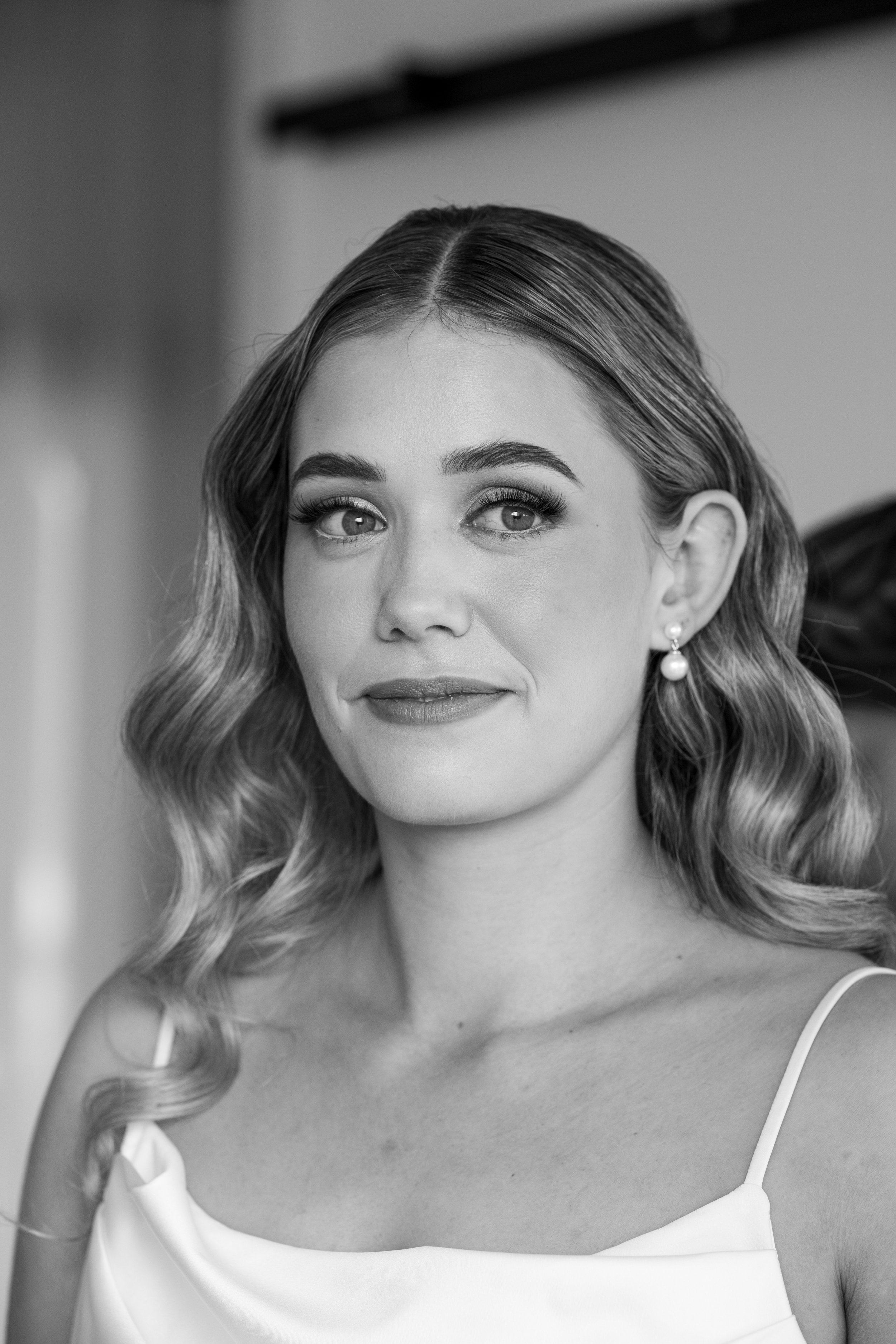 A black-and-white portrait of a woman with wavy hair, wearing a sleeveless top and pearl earrings.