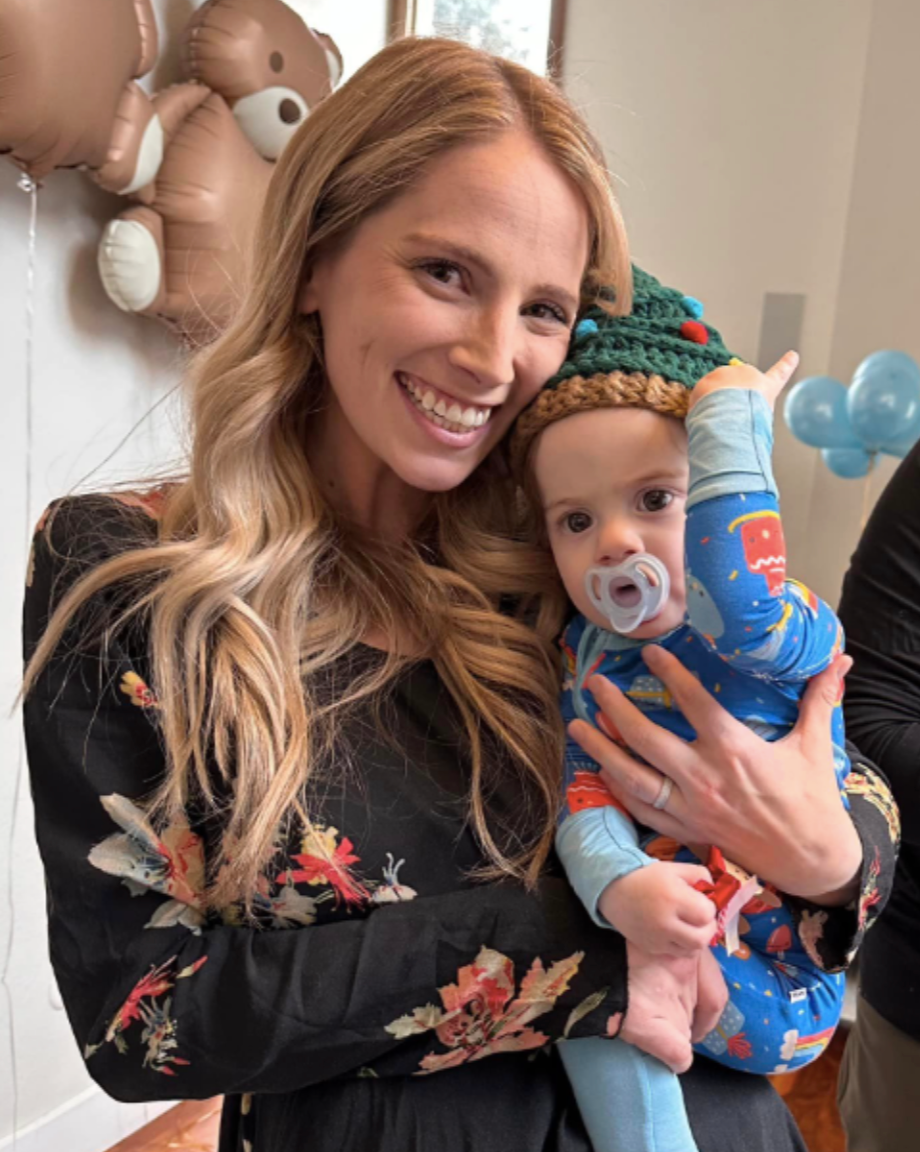 A smiling woman with long blonde hair holding a baby wearing a colorful elf hat and blue pajamas with festive patterns, in a decorated room with balloons and teddy bear-shaped balloons in the background.