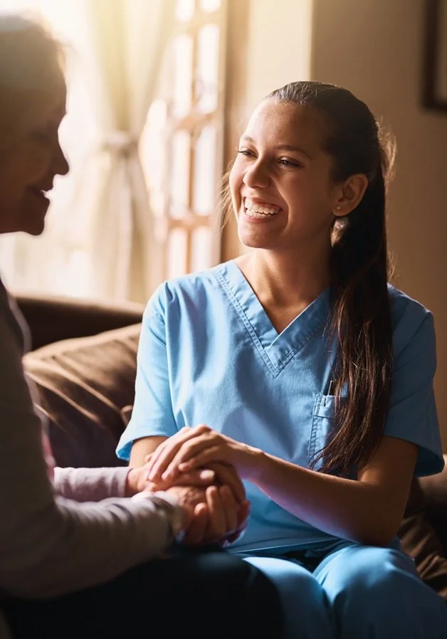 A young woman in medical scrubs smiling and holding hands with an older woman in a cozy room with sunlight streaming through the window.
