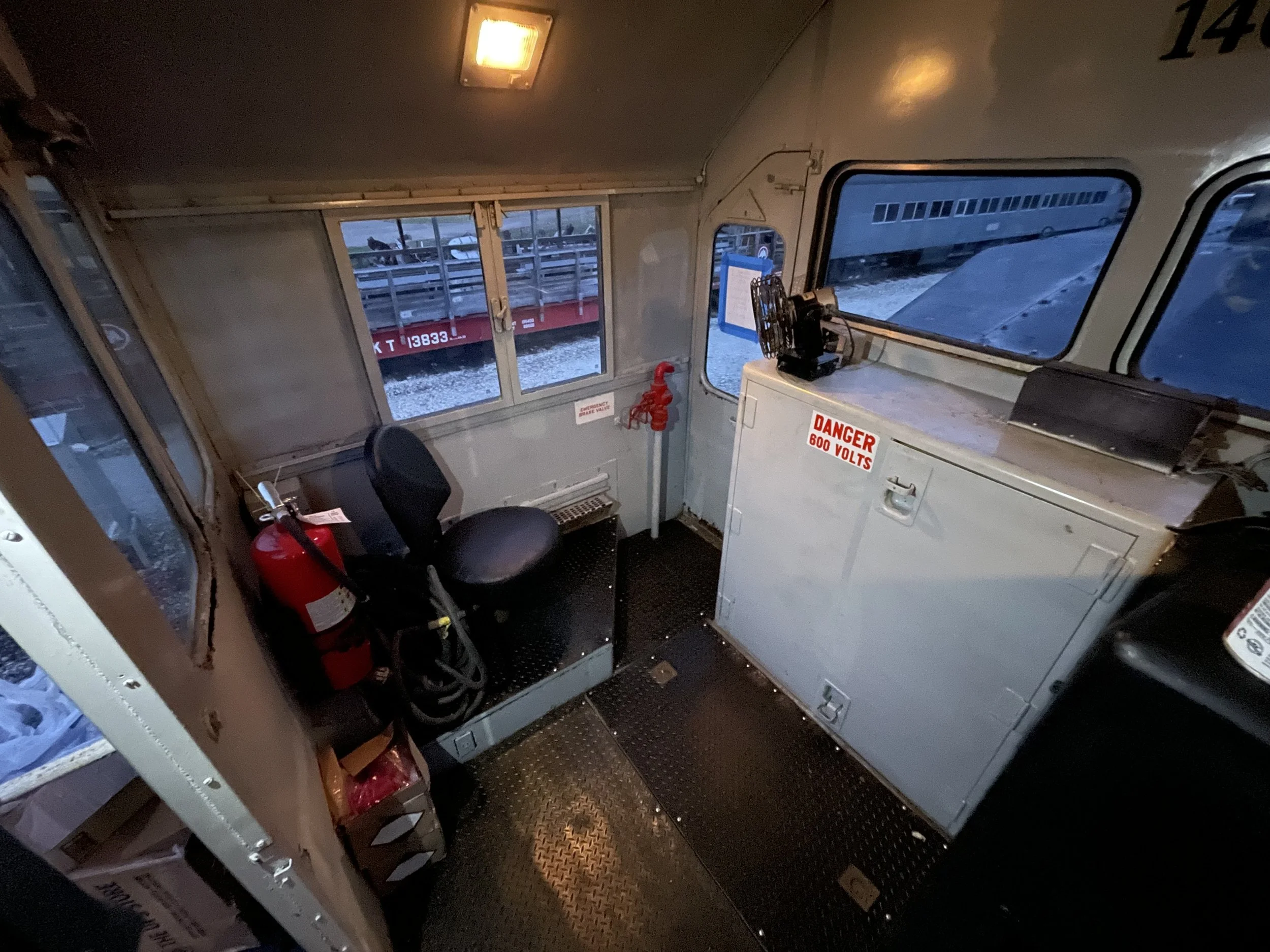 Interior of a locomotive cab with controls, gauges, and a fan, view through windows showing train yard.