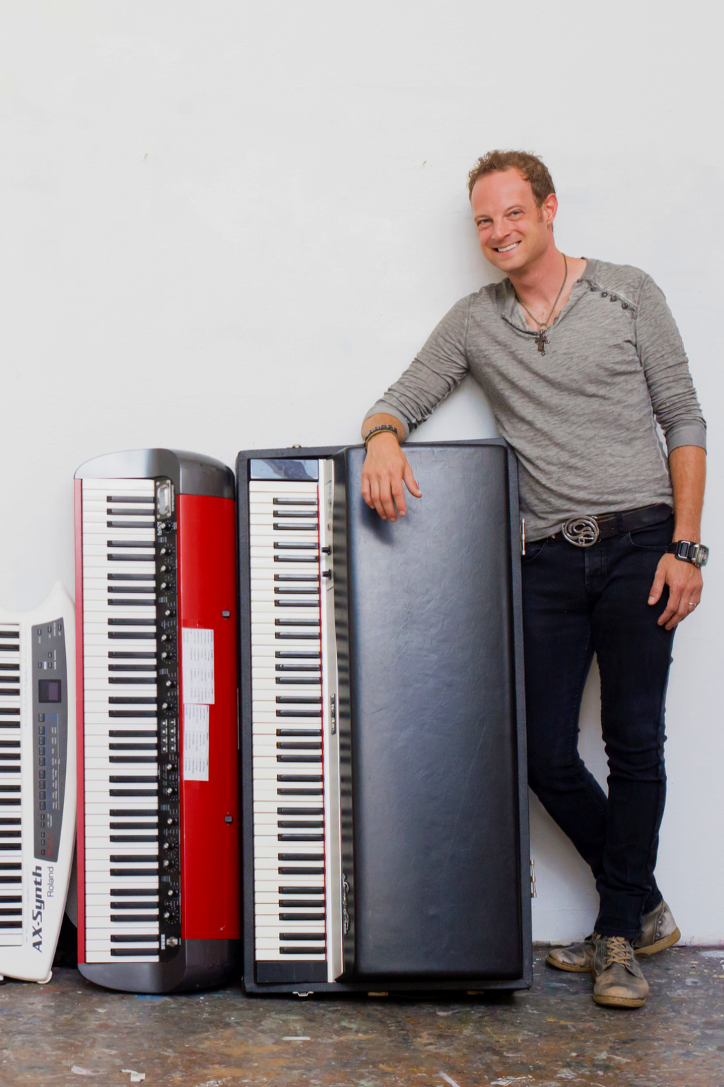 A man in a gray long sleeve shirt and black pants leaning on a piano, with several electronic keyboards and a black case nearby, standing against a white wall.