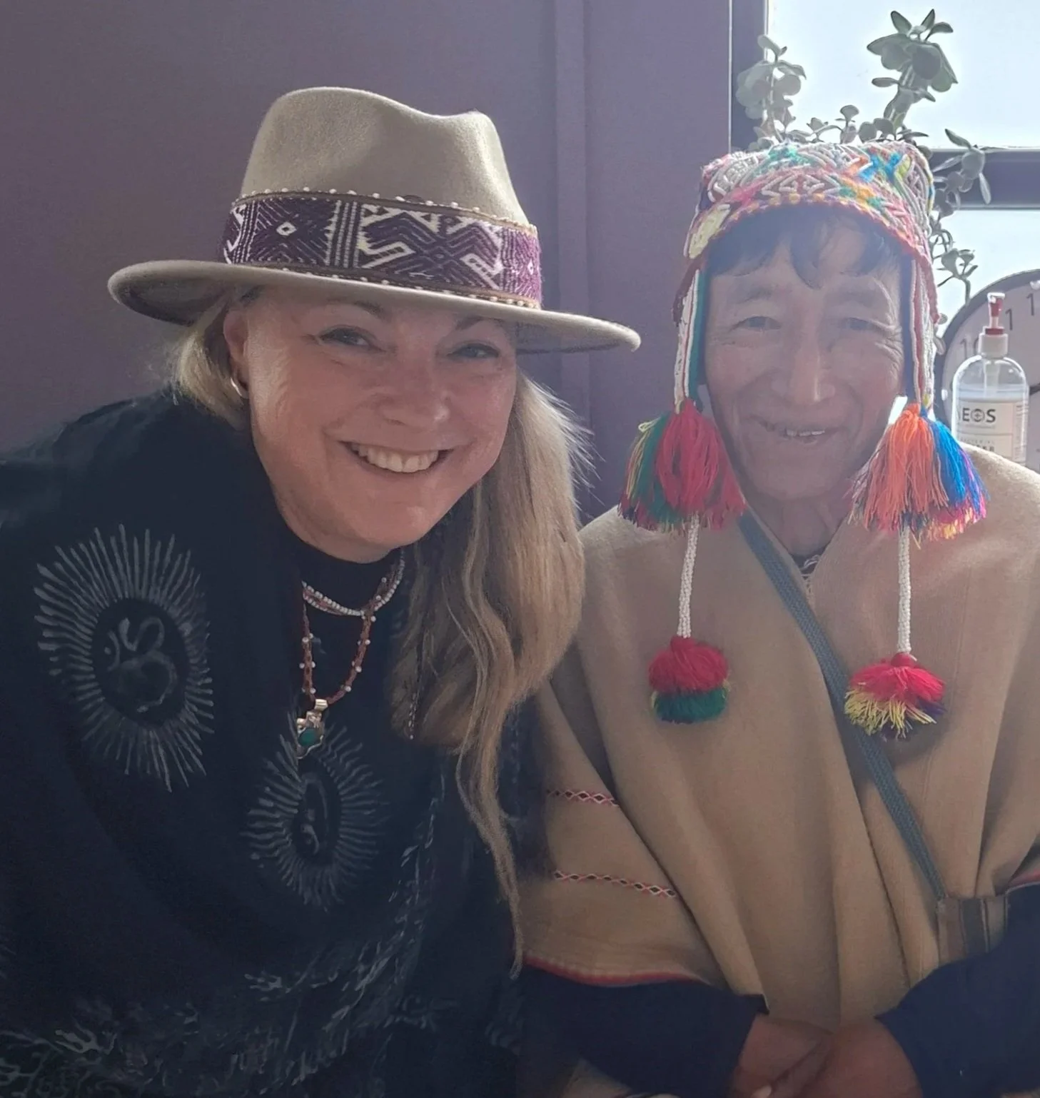 Two women smiling, one wearing a beige felt hat with a colorful band and the other wearing a traditional colorful woven hat with tassels. They are sitting indoors near a window with plants in the background.