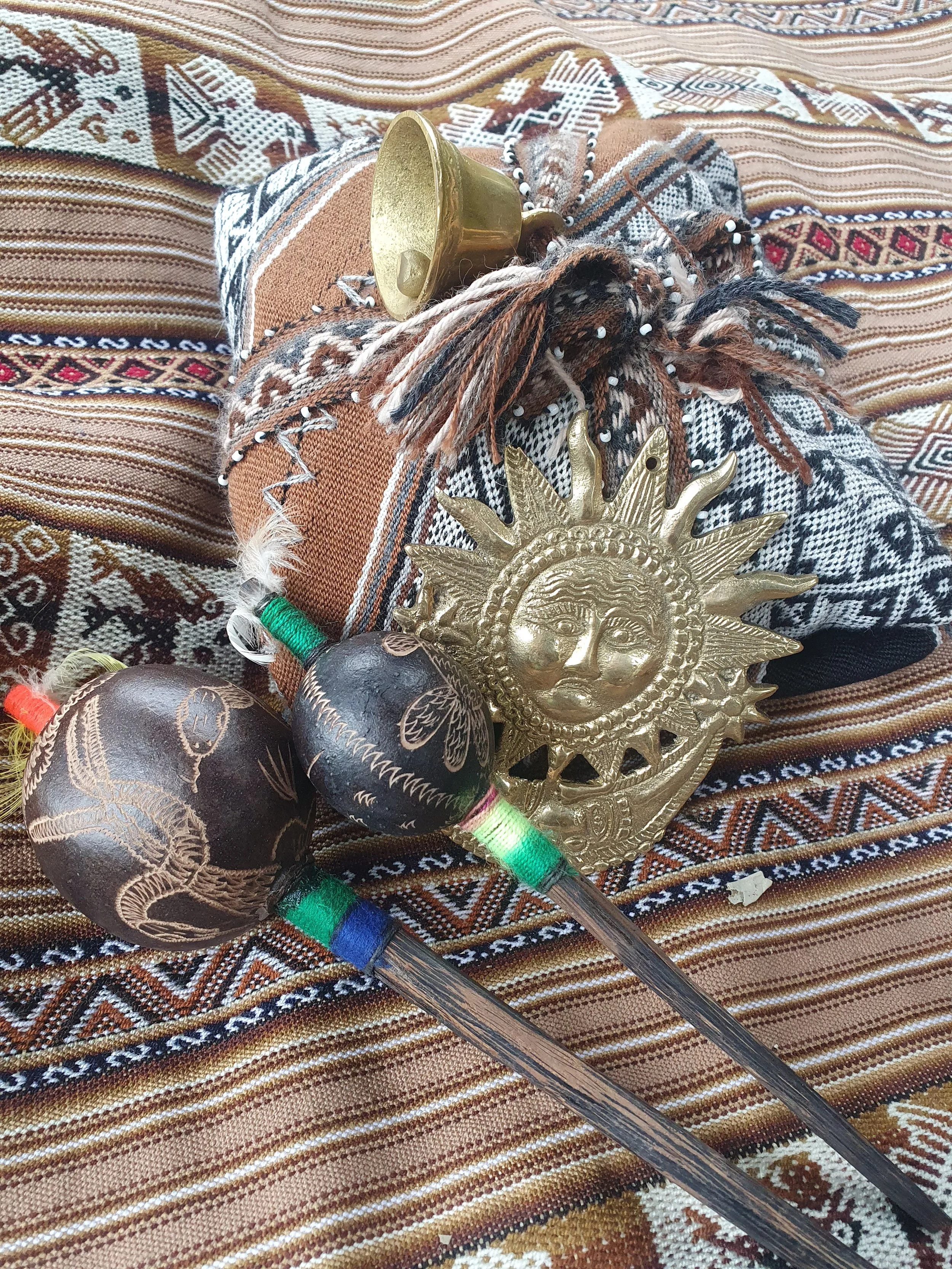 A peruvian woven tapestry with a medicine bundle and 2 shamanic rattles next to a golden decorated sun-shaped medal.