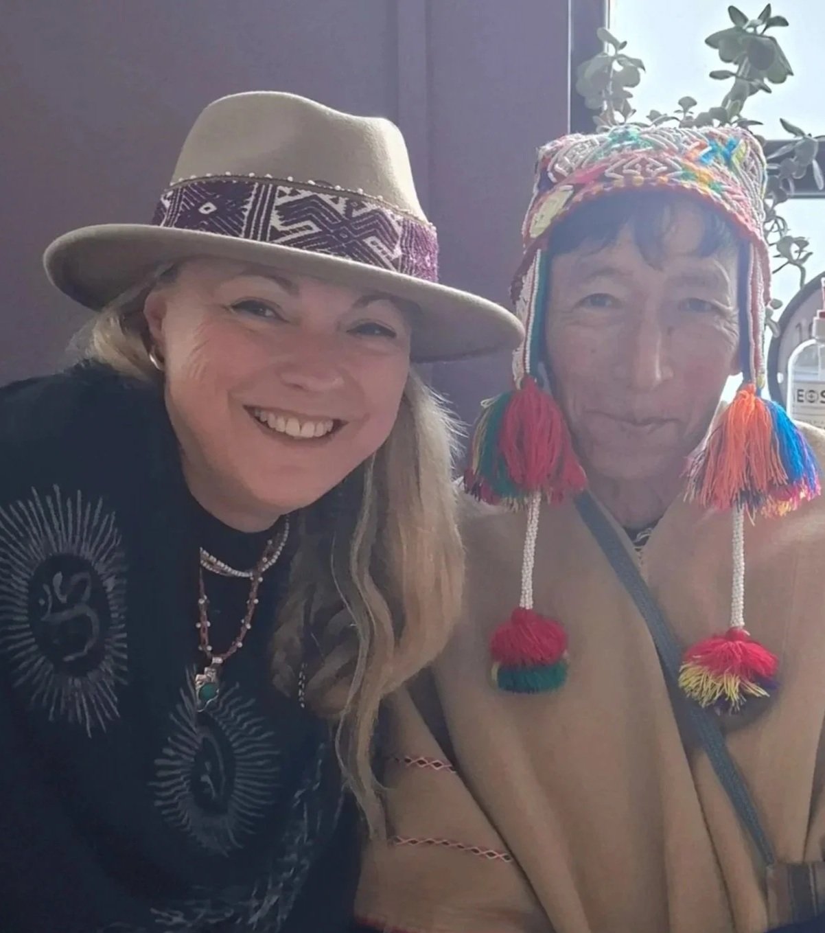 Two women wearing hats and traditional attire, smiling for a selfie indoors with a plant and sunlight in the background.
