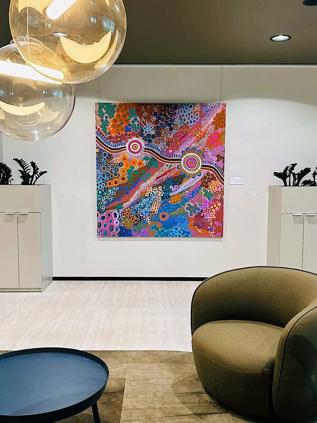 Modern interior with abstract, colorful artwork on the wall, a beige armchair, a small blue table, and decorative plants in white planters.