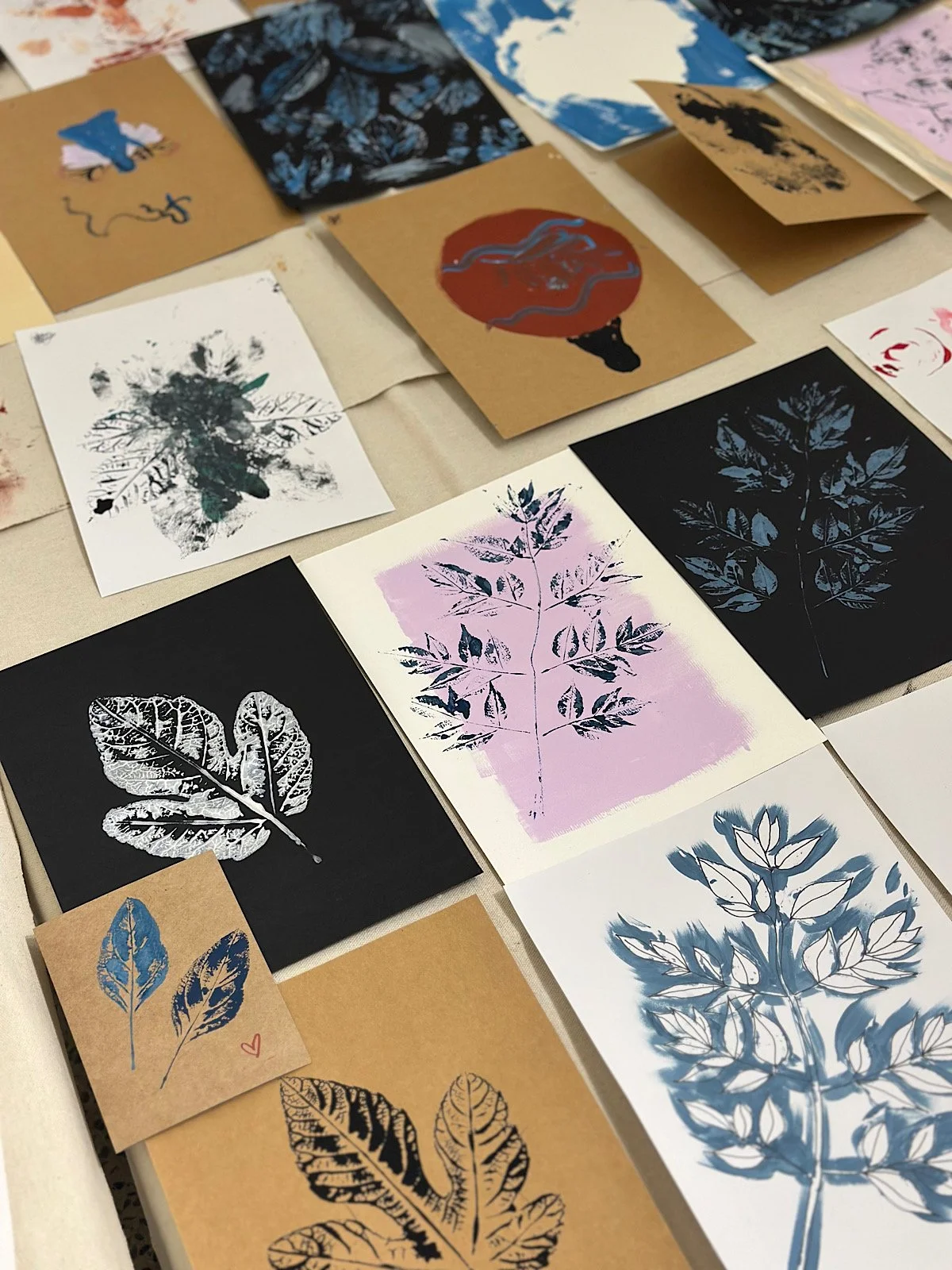 Various art prints and postcards featuring leaf and plant silhouettes, some with watercolor effects, in black, white, blue, purple, and pink, arranged on a table.
