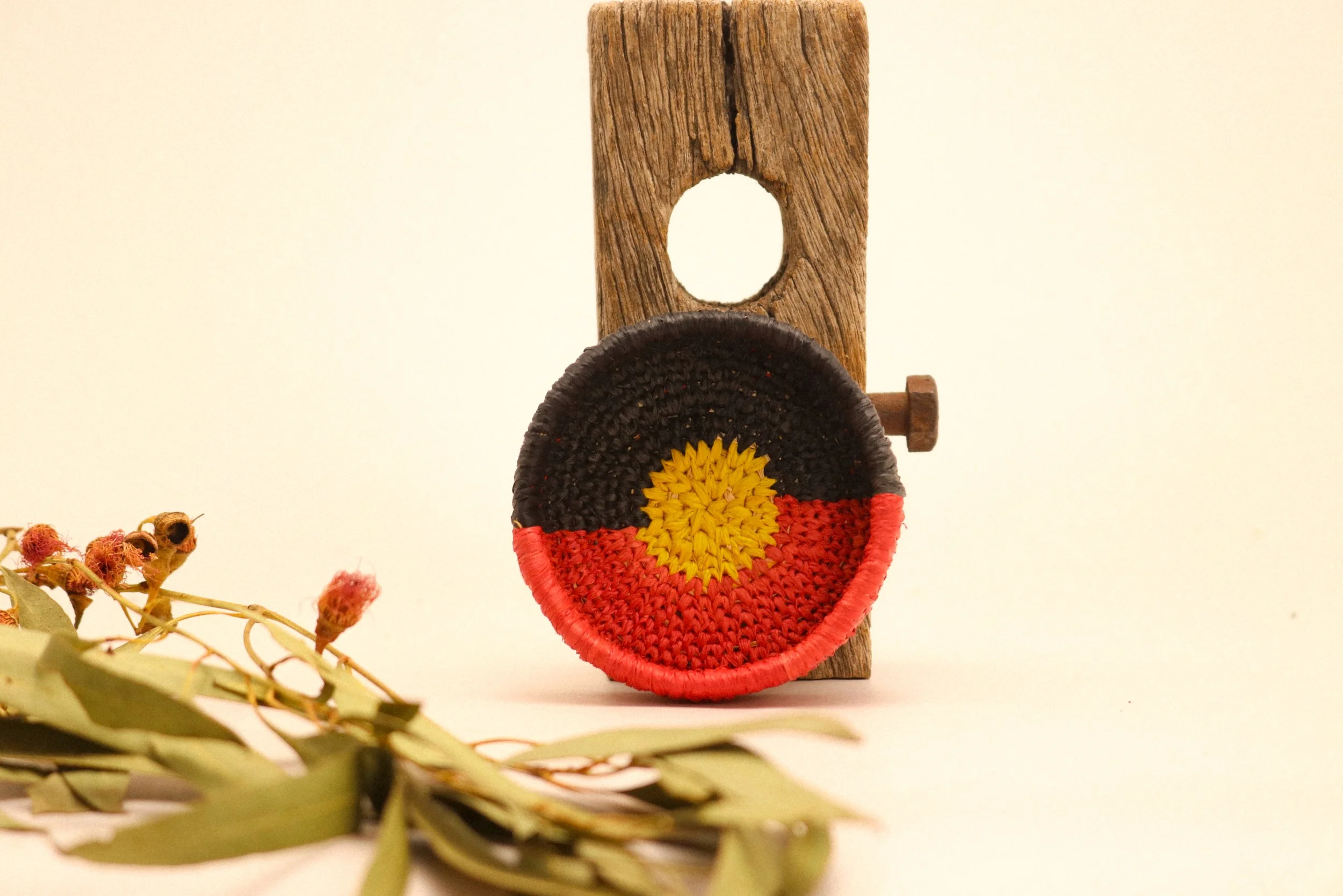 A small woven circular item with the colors of the Australian flag, positioned behind a wooden tool with a hole at the top, next to a bunch of dried leaves and small flower buds.