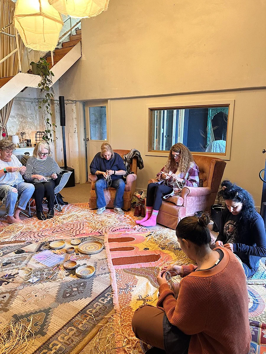 A group of women knitting and working on craft projects together in a cozy room with an ornate rug, armchairs, and decorative lighting.