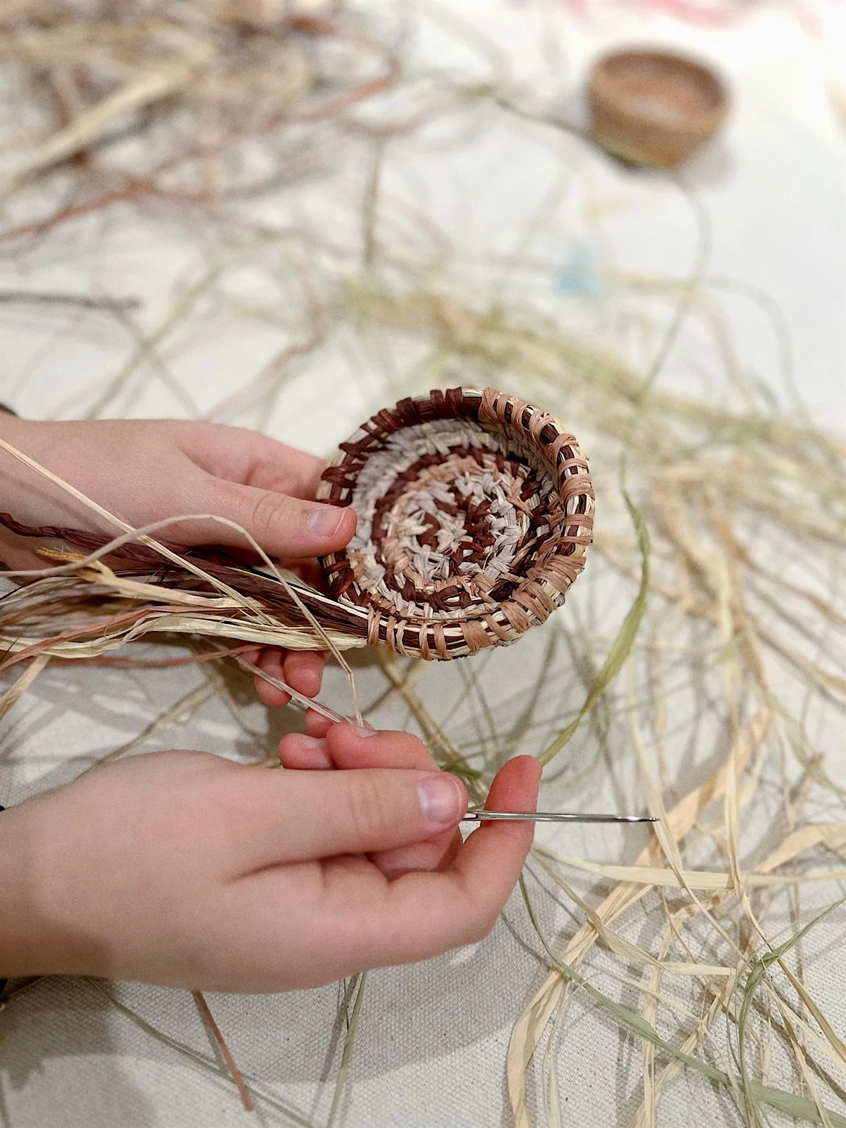 A person weaving a small basket using natural fibers, with a needle, surrounded by dried grass and fibers on a neutral background.