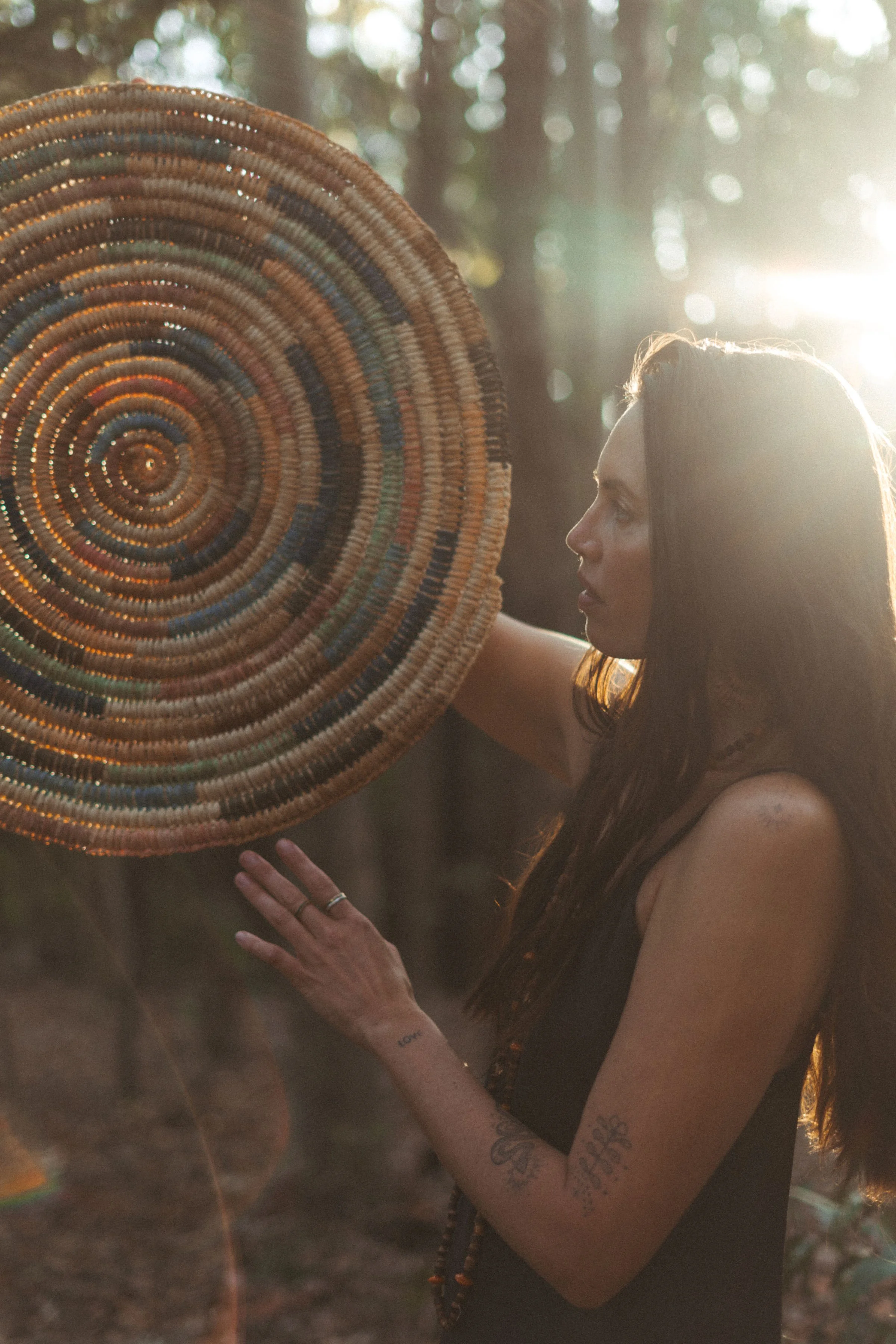 Woman with long dark hair holding a large, woven, circular basket in a forest setting during sunset.