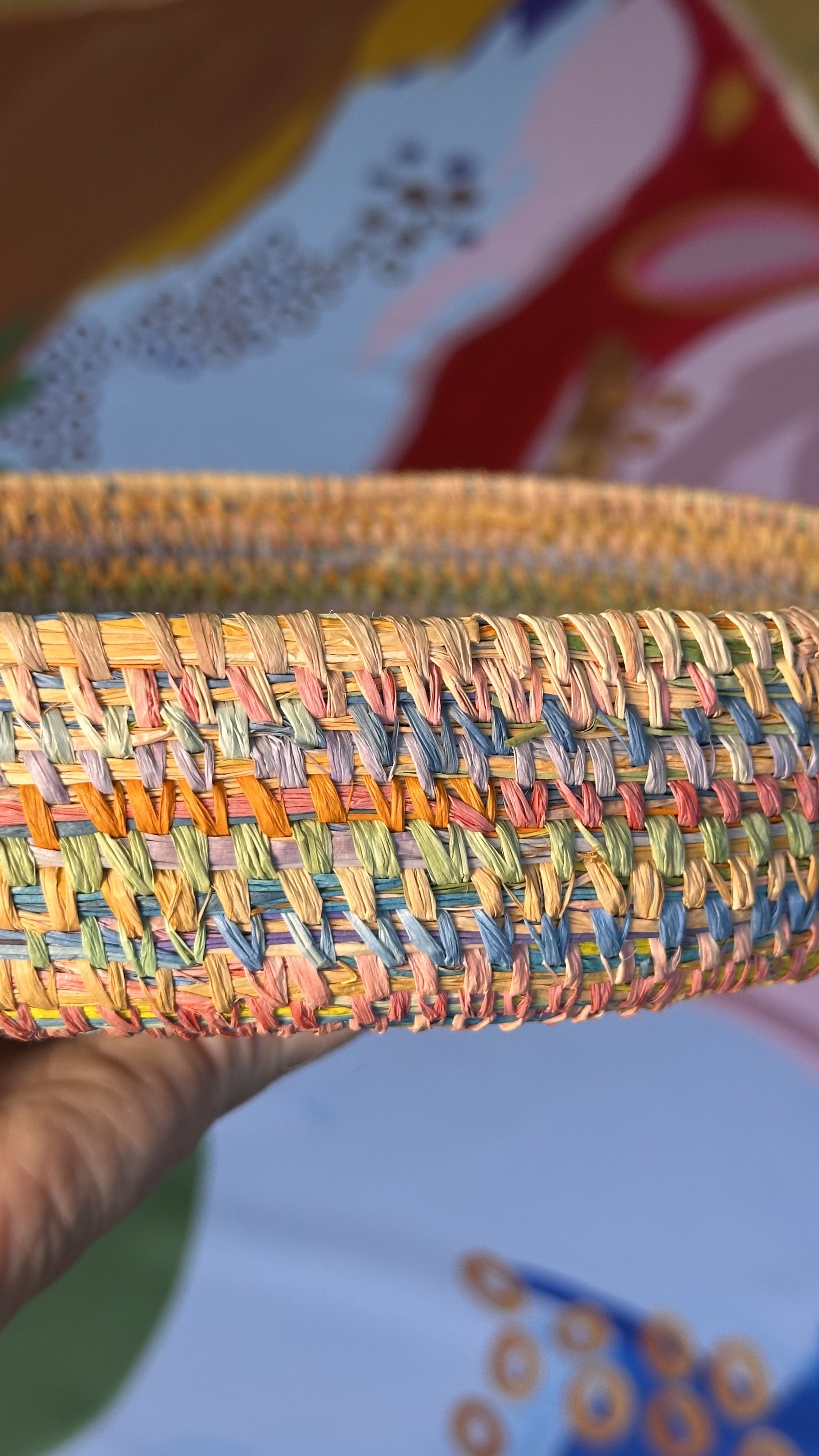 Close-up of a woven multicolored basket made from straw or similar material, held in a person's hand with a blurred background.