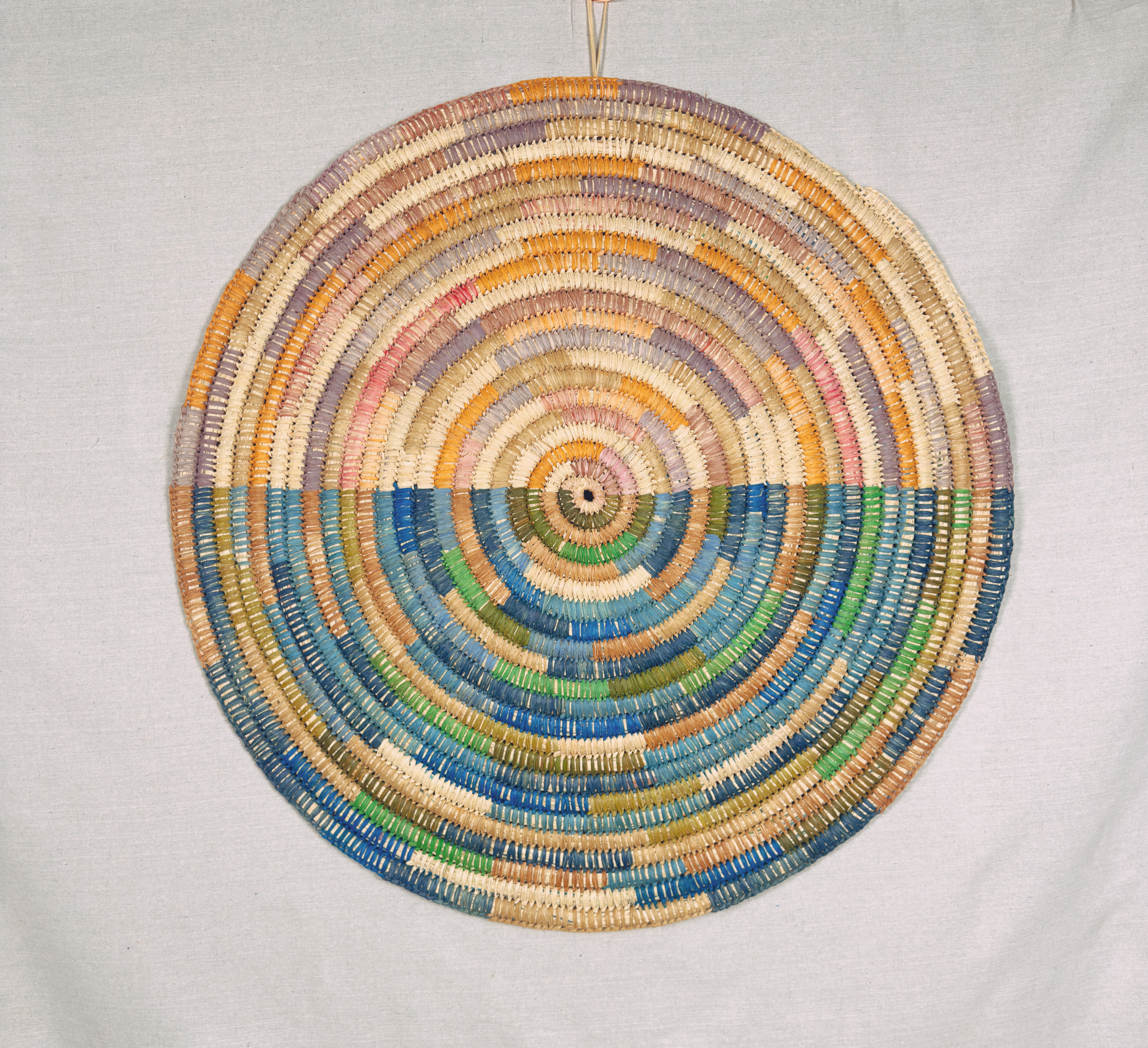 Colorful woven circular wall art with rainbow pattern on plain white background.