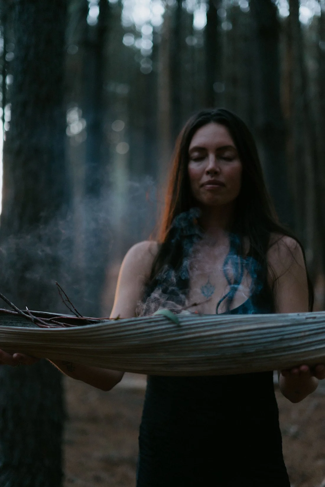 A woman with long dark hair stands in a forest, holding a large wooden bowl with smoke rising from it, and appears to be in a meditative or ritual pose.