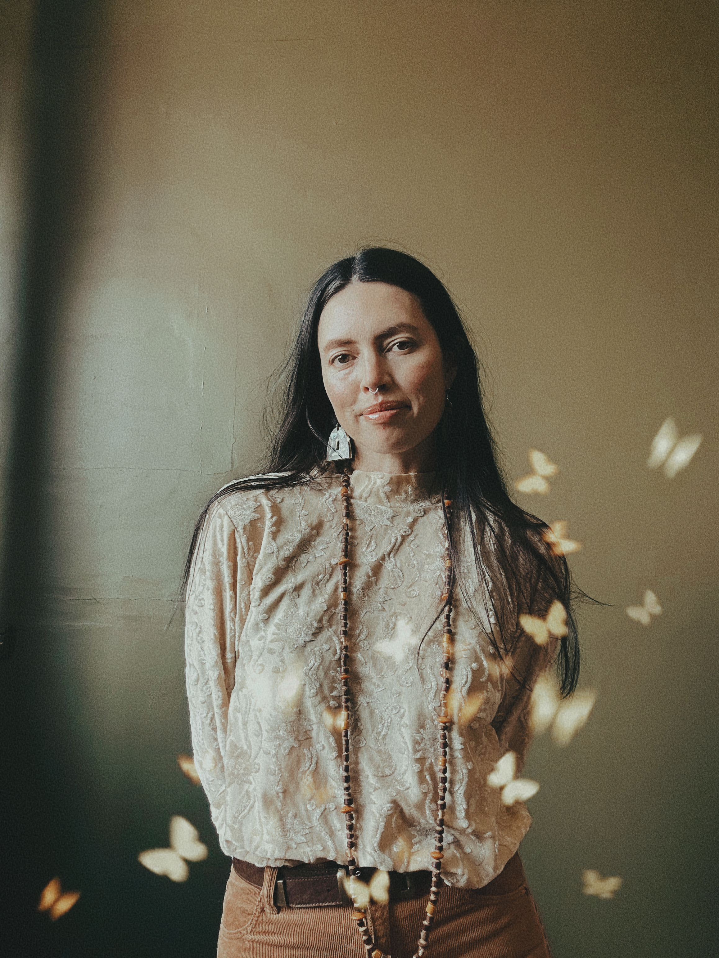A woman with long dark hair, wearing a beige patterned blouse, brown pants, large earrings, and a beaded necklace, standing indoors with a plain wall background, surrounded by floating butterfly effects.