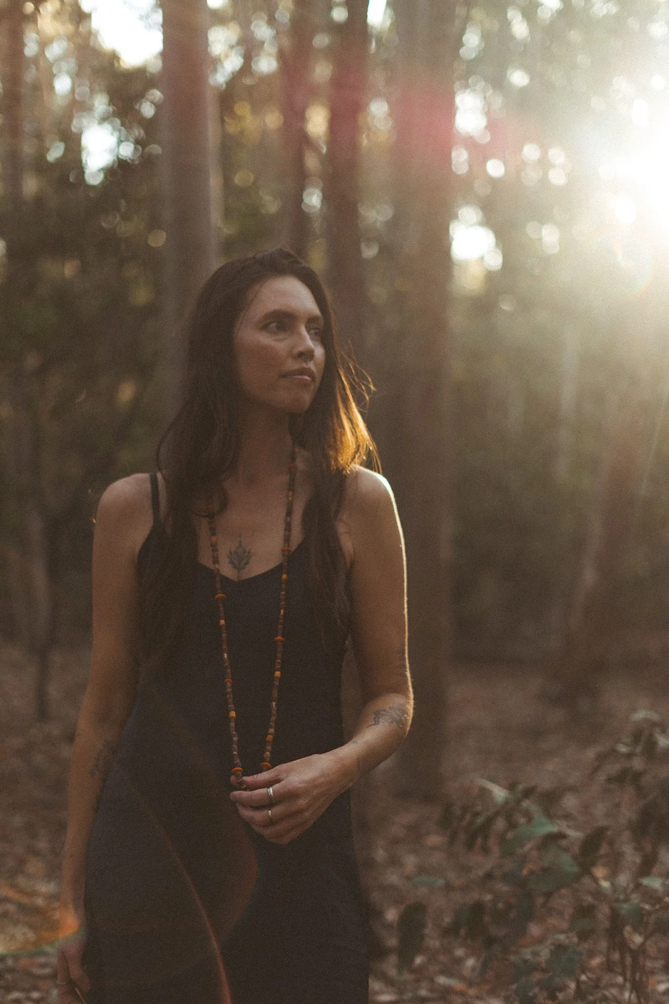 A woman with long dark hair wearing a black dress and a beaded necklace stands outdoors in a forest, illuminated by warm sunlight.