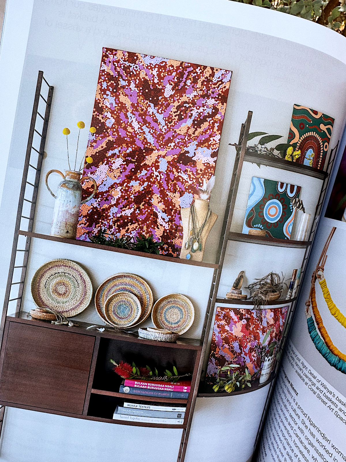 A display of colorful artwork and handcrafted items on a multi-tiered shelving unit. The top shelf features a large abstract painting with pink, purple, blue, and brown colors, along with a ceramic jug holding yellow globular flowers and a mannequin bust with jewelry. The middle shelf holds smaller patterned baskets. The shelves below contain books, a small plant, and additional artwork.