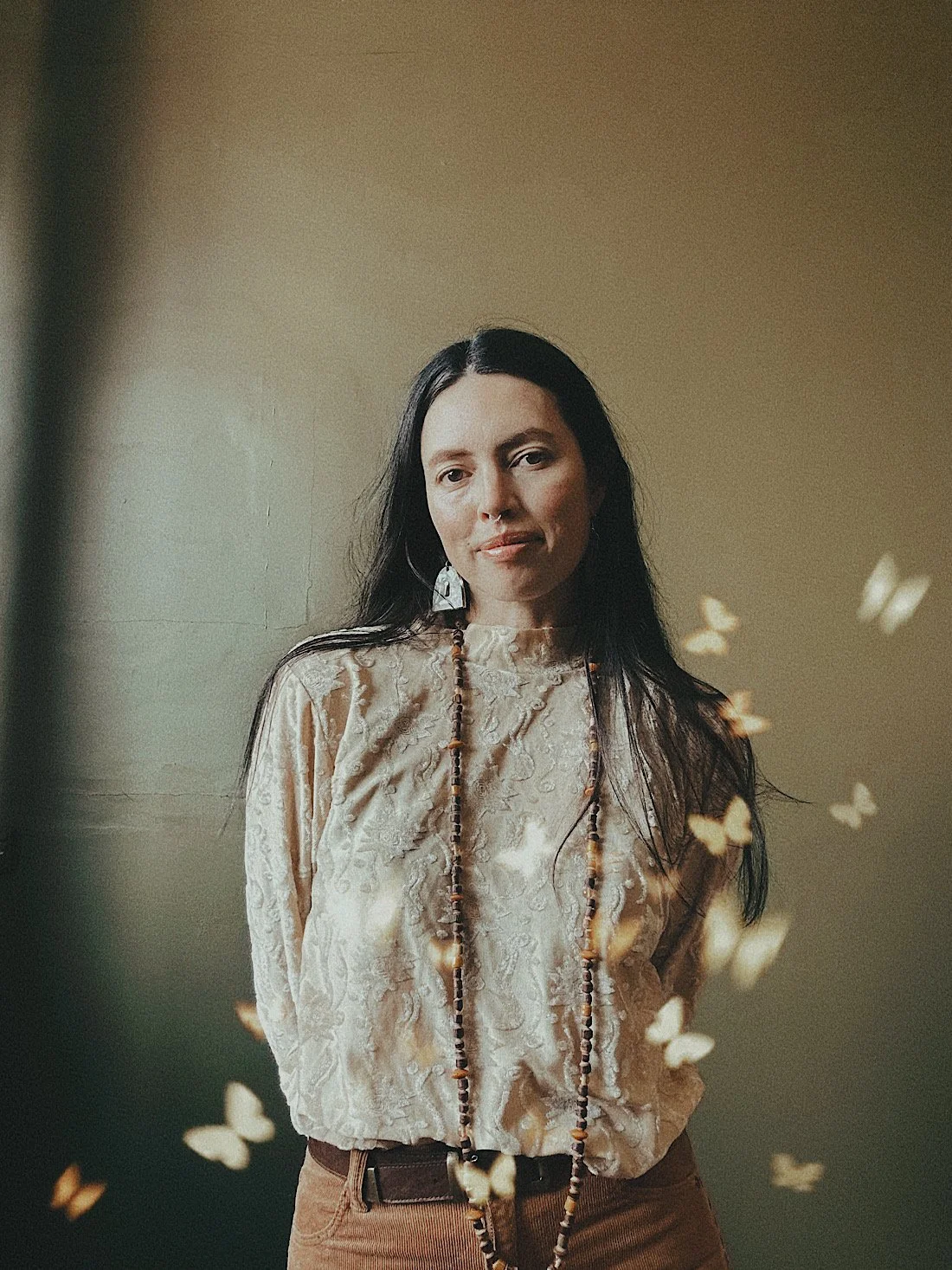A woman with long dark hair wearing a beige, patterned blouse and beaded necklace. She is standing against a plain, light-colored wall with soft light and small butterfly illustrations floating around her.