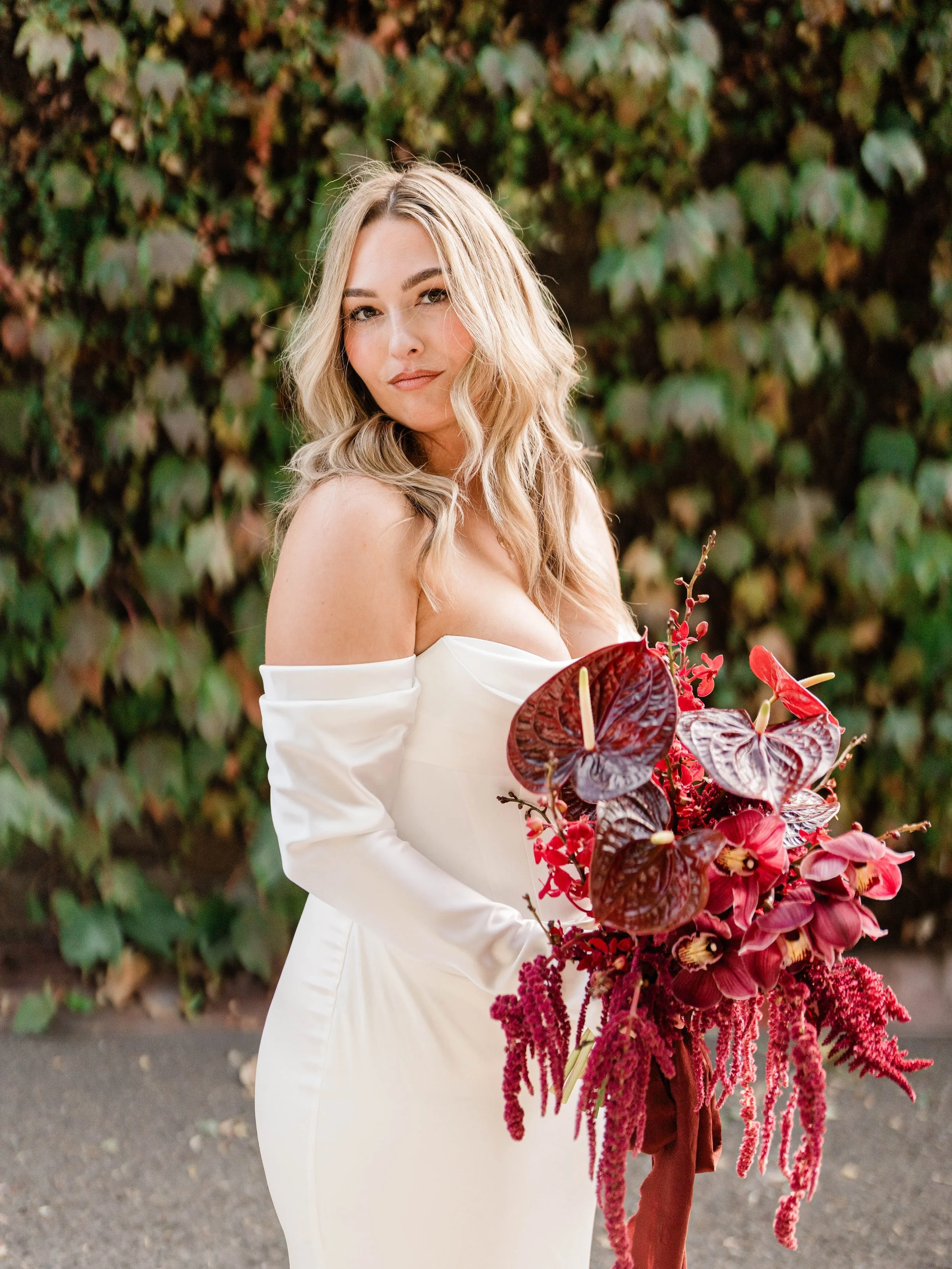 Bride holding a deep red bouquet with anthuriums and orchids in front of green ivy wall.