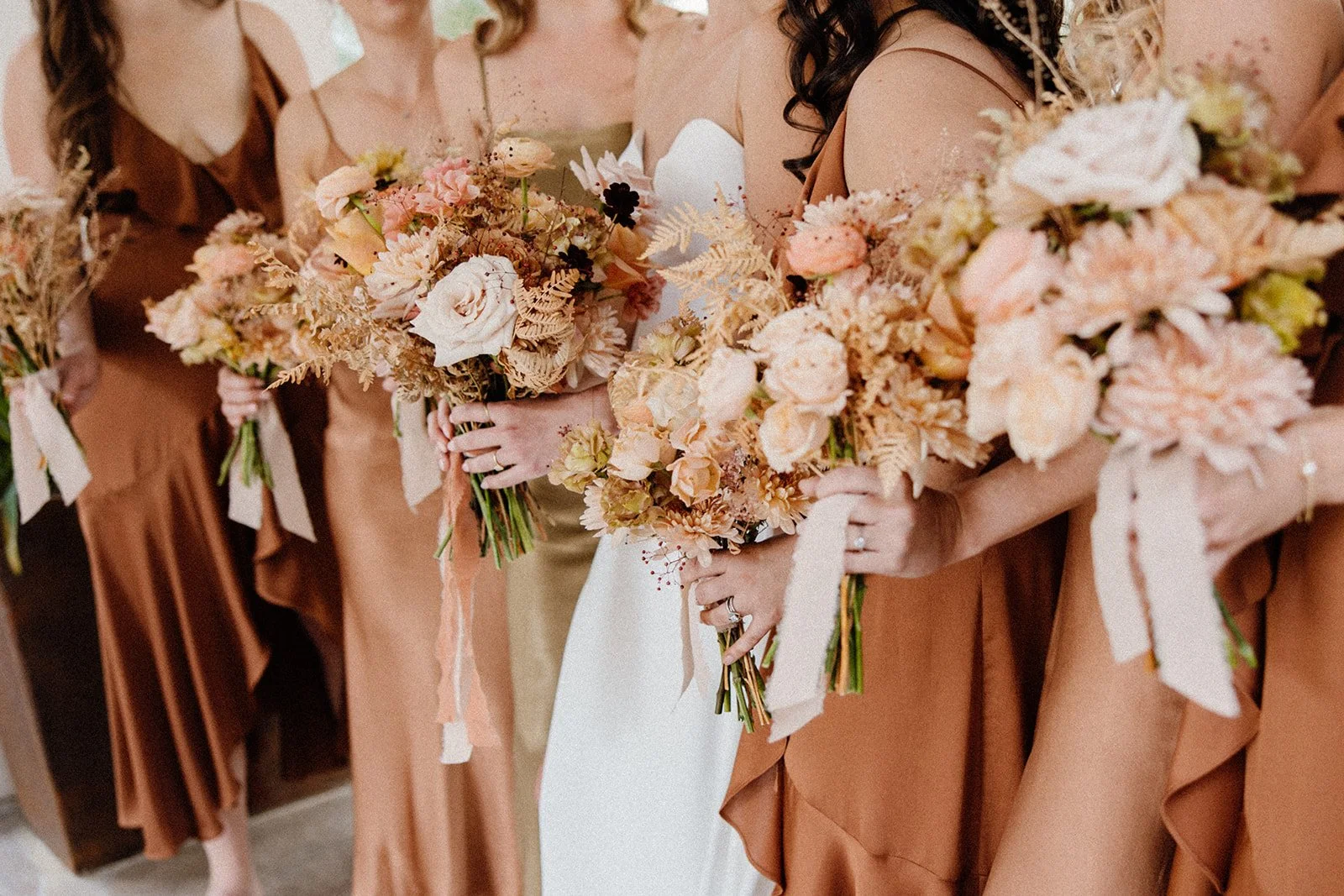  Close-up of bridesmaid and bridal bouquets featuring blush roses, soft peach blooms, dried textures, and trailing ribbons for a fall wedding at La Quinta Resort. 
